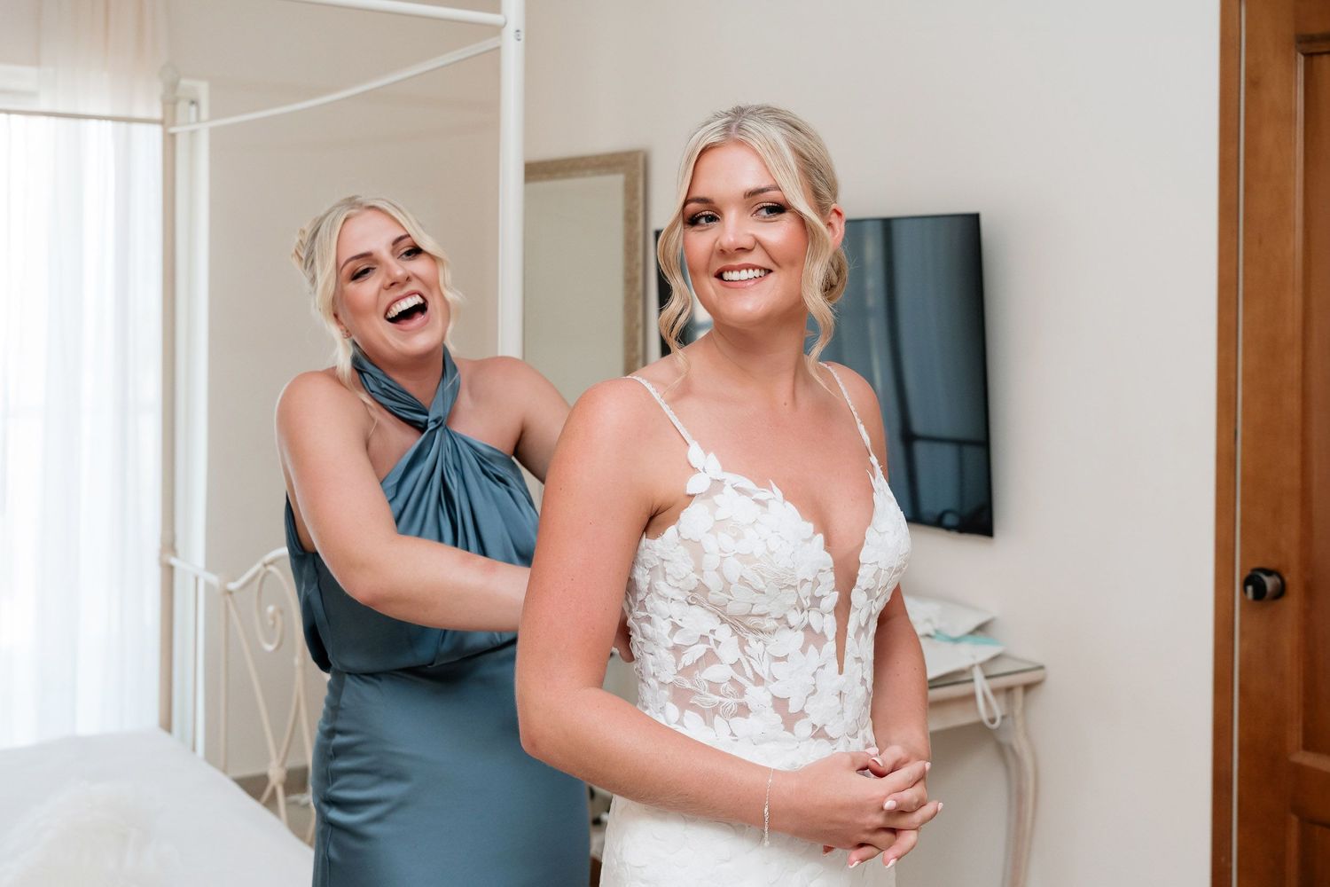 Two people share a joyful moment while getting ready in a bright room with a white dress.