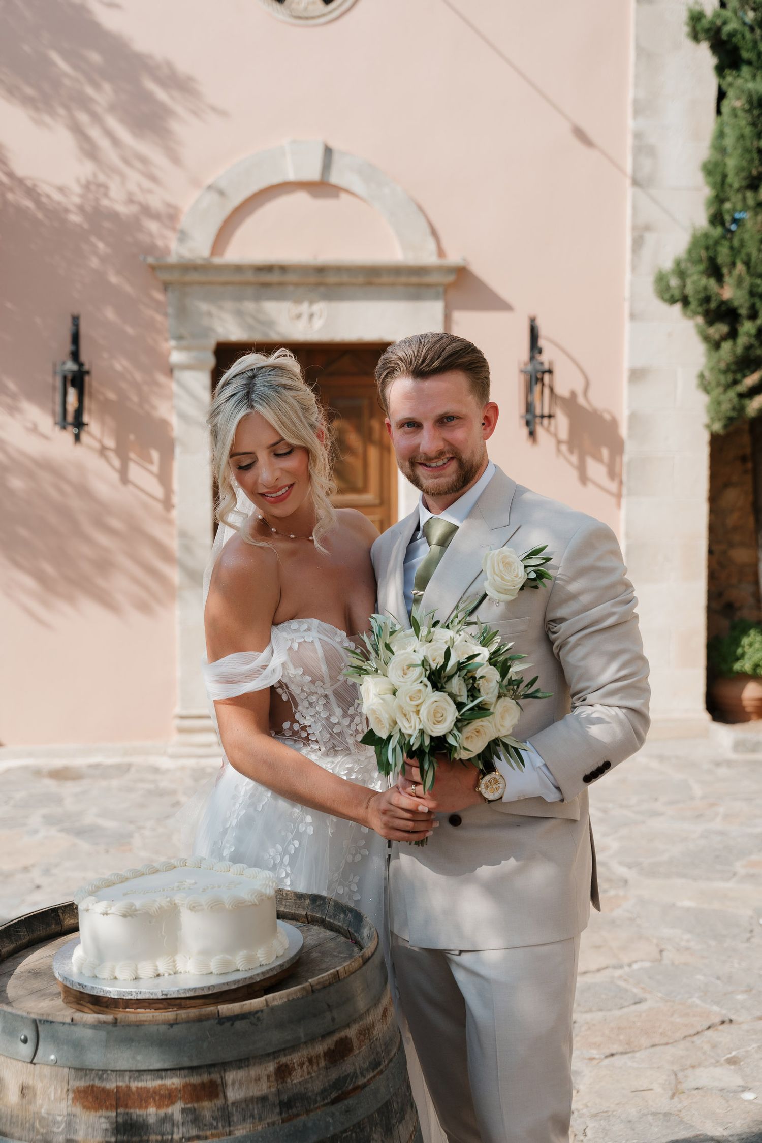 A newlywed couple in light-colored attire poses with a white floral bouquet next to a rustic wine barrel and pink wall.