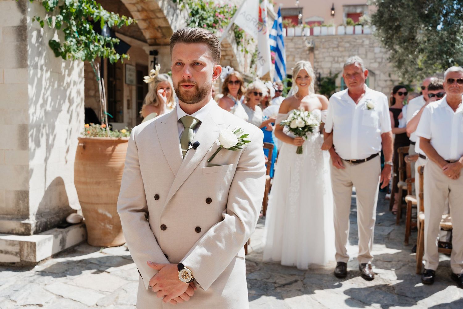 A wedding party gathers in a Mediterranean courtyard with stone walls and greenery overhead during a sunny celebration.