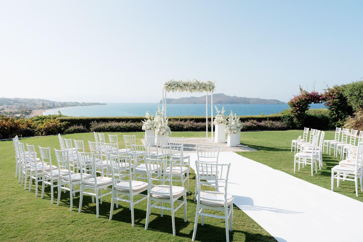 White wedding chairs arranged in rows on a grassy lawn overlooking a scenic ocean view for an outdoor ceremony.
