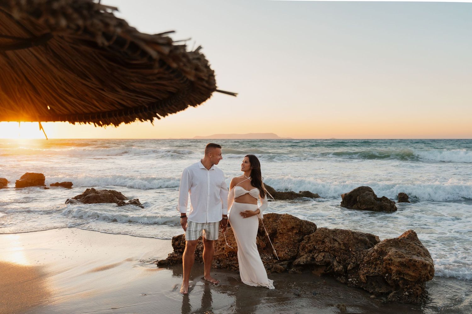 Romantic coastal sunset photoshoot on a rocky beach with crashing waves and golden light.