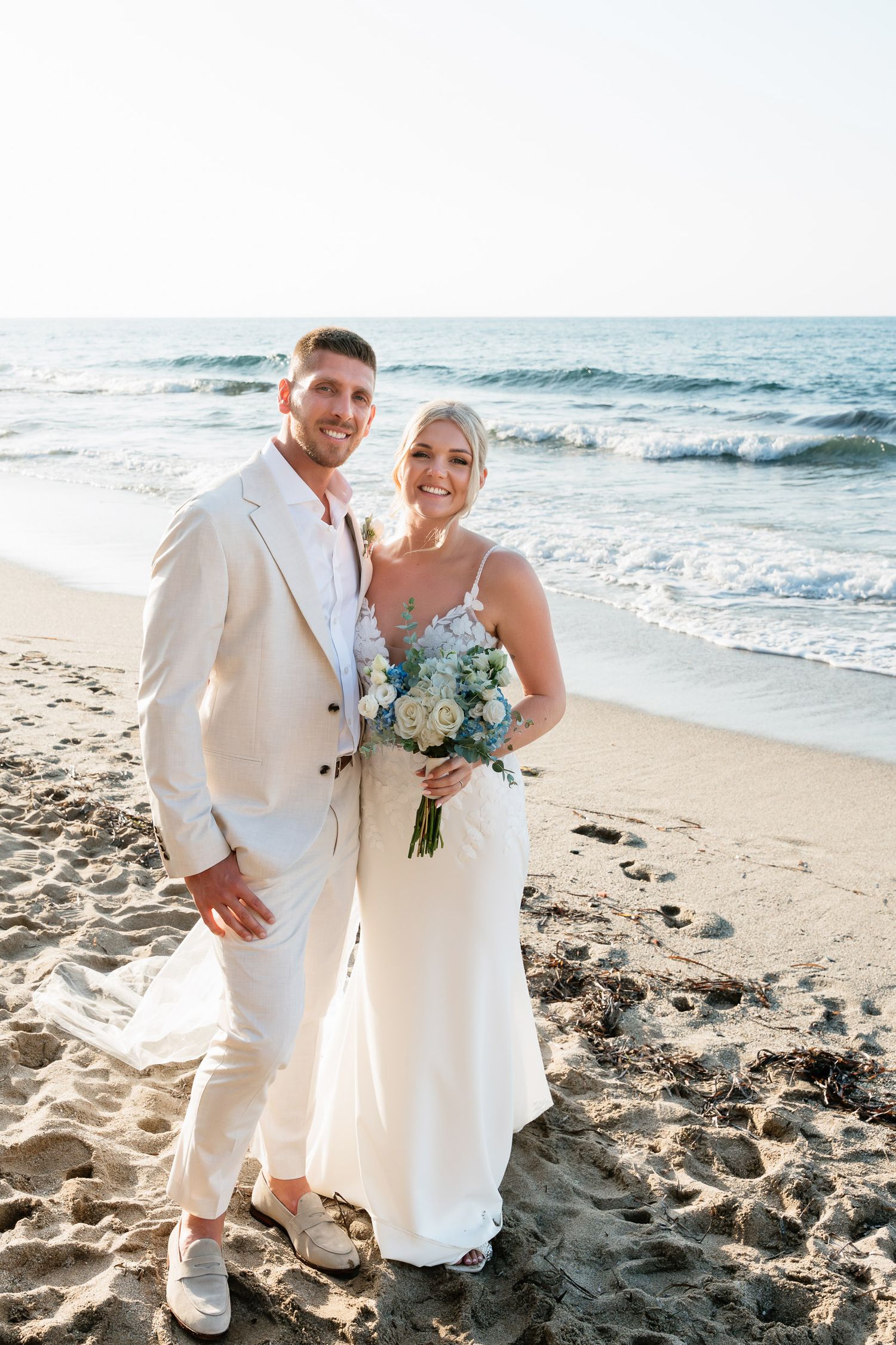 A couple poses together on a sunny beach with ocean waves in the background during their wedding celebration.