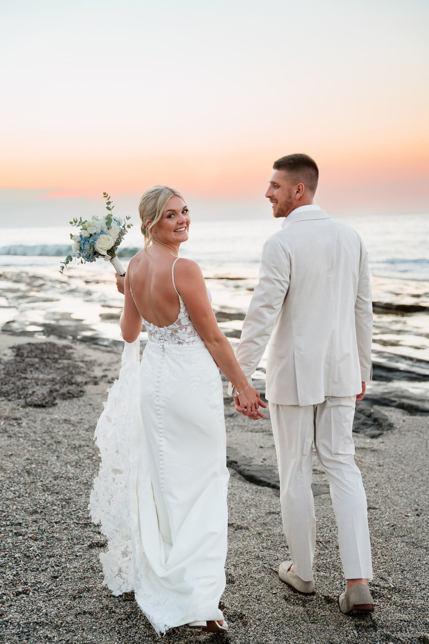 A couple in wedding attire walks along a rocky beach at sunset, holding hands and enjoying the romantic coastal scenery.
