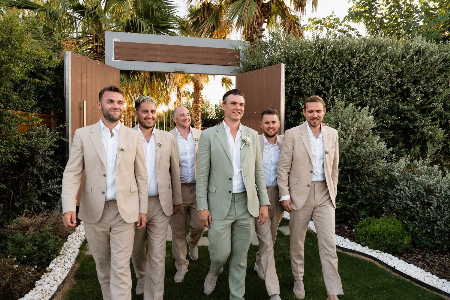 Wedding party poses together in light beige and sage green suits at an outdoor venue during golden hour.