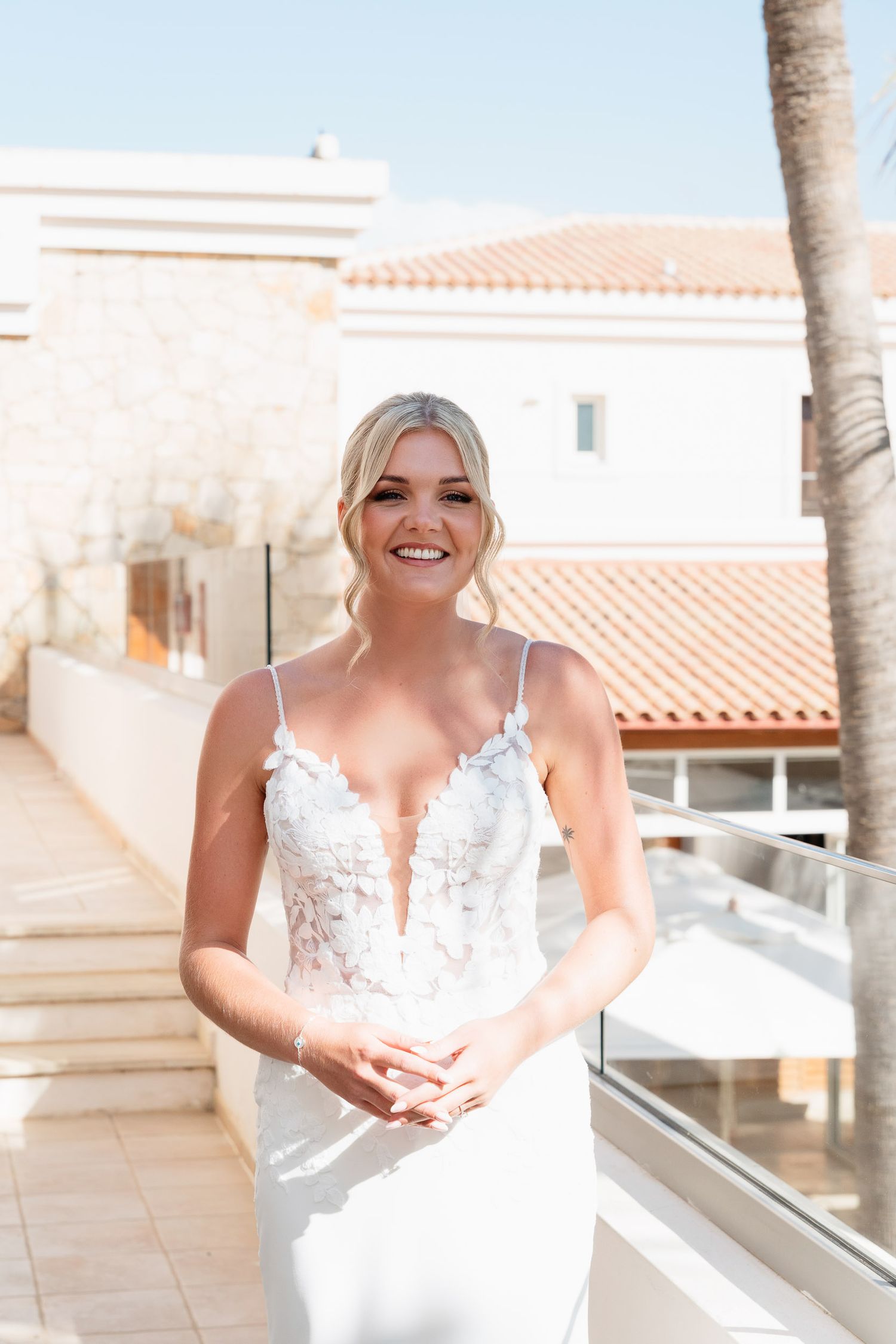 A bride in a white lace wedding dress sits on a balcony with terra cotta roofs and palm trees in the background.