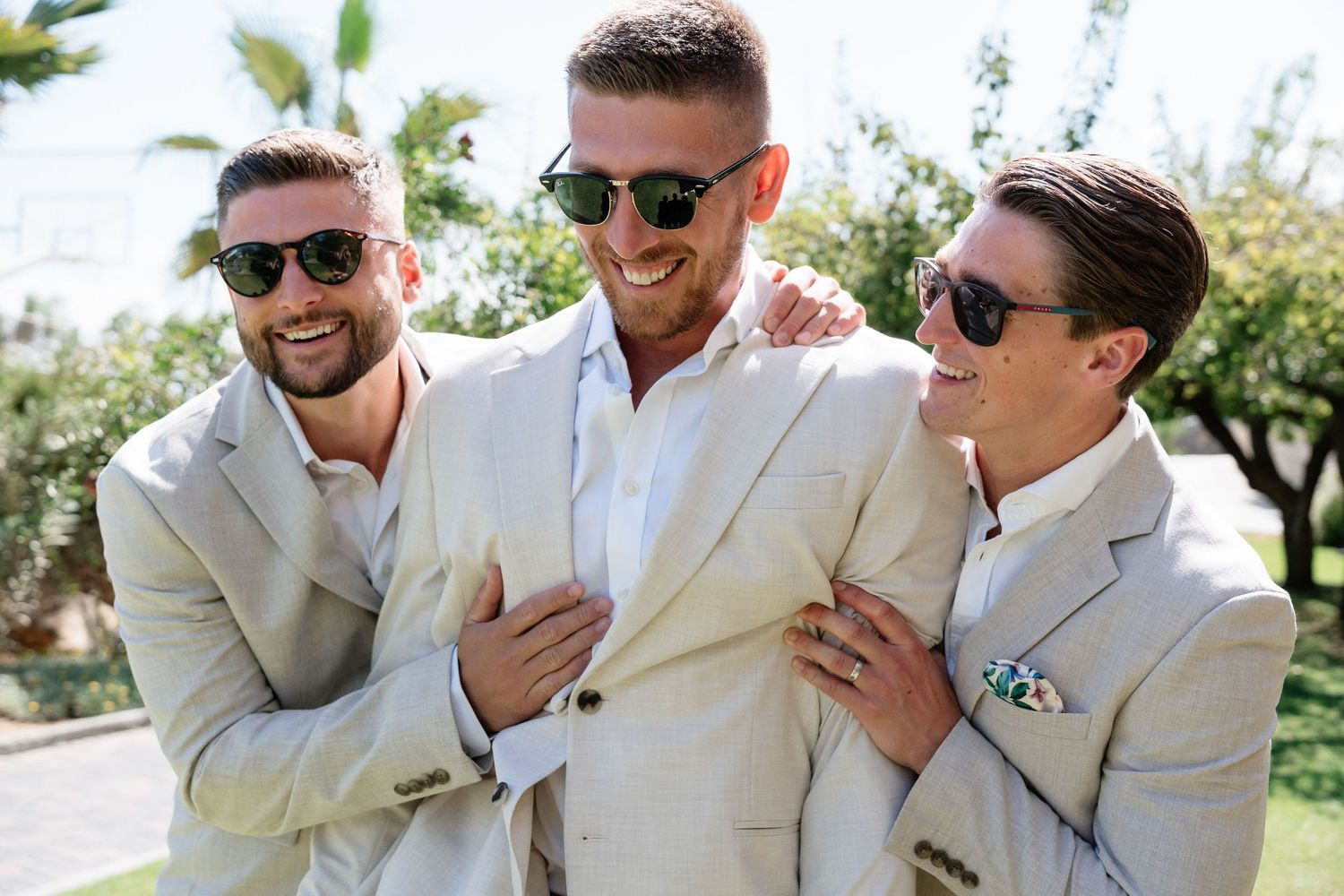 Three people in light colored suits and sunglasses laugh together outdoors on a sunny day with palm trees in background.