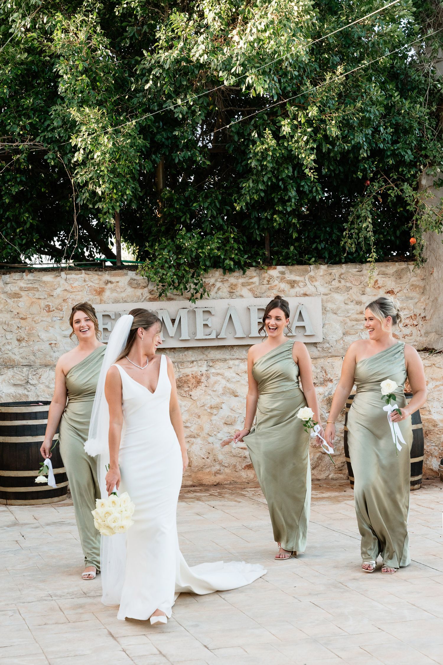 Wedding party in sage green dresses and white gown walking together at an outdoor venue with stone walls.