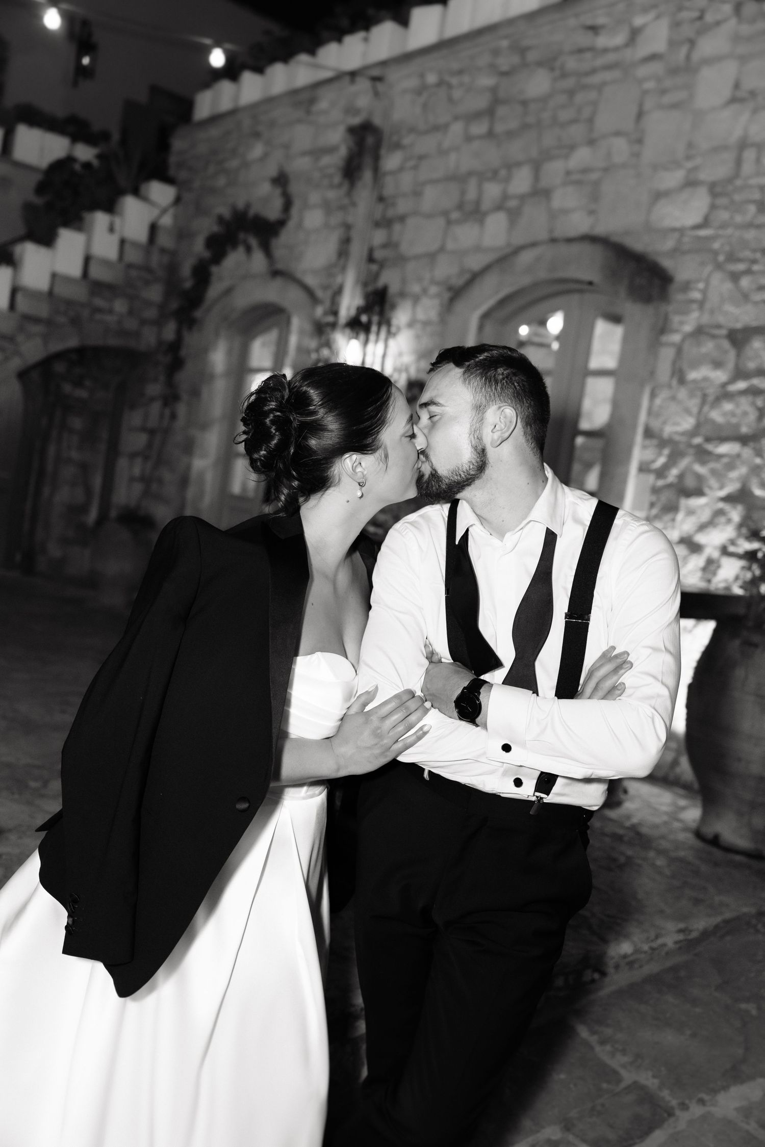 Black and white wedding photo of bride and groom sharing a kiss outdoors at a stone venue at night.