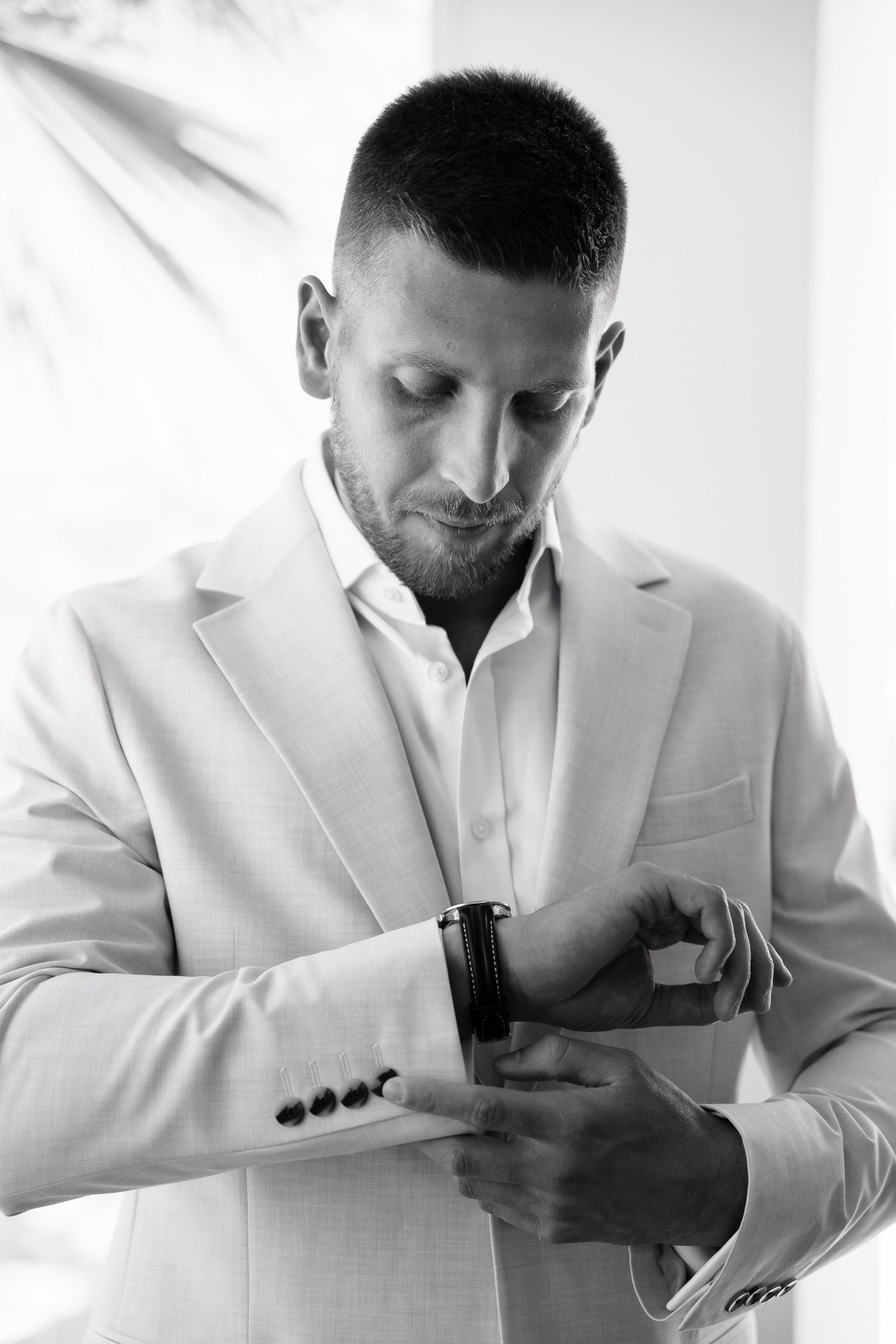 A man in a white suit jacket checks his watch while standing against a light background in black and white photography.
