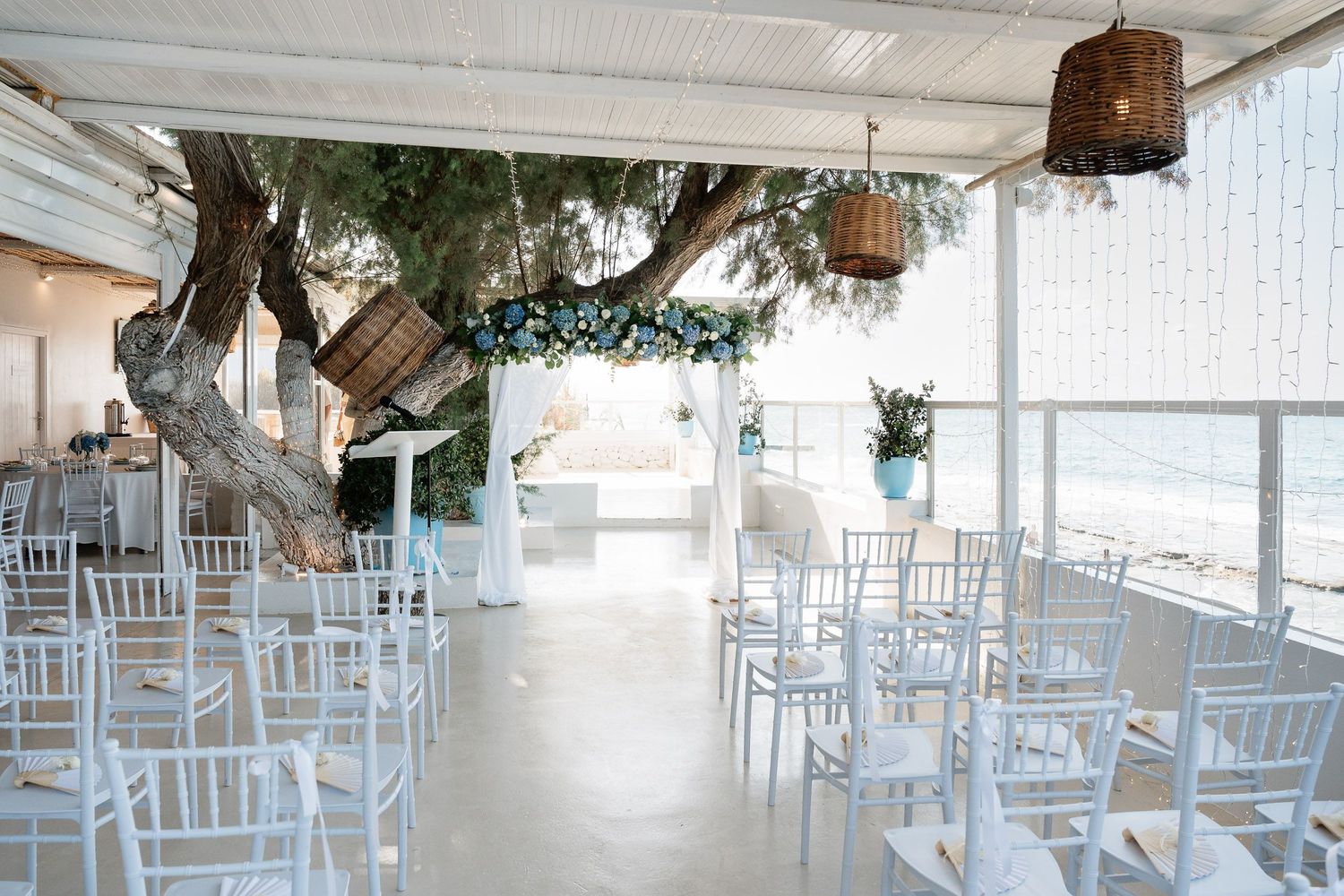 White wedding reception venue setup with rows of chairs and floral decorations in an airy space.