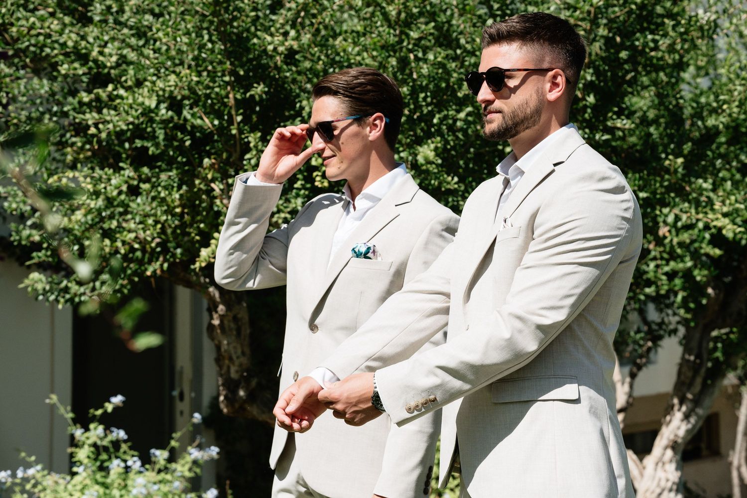 Two people in matching light colored suits and sunglasses stand together outdoors on a sunny day.