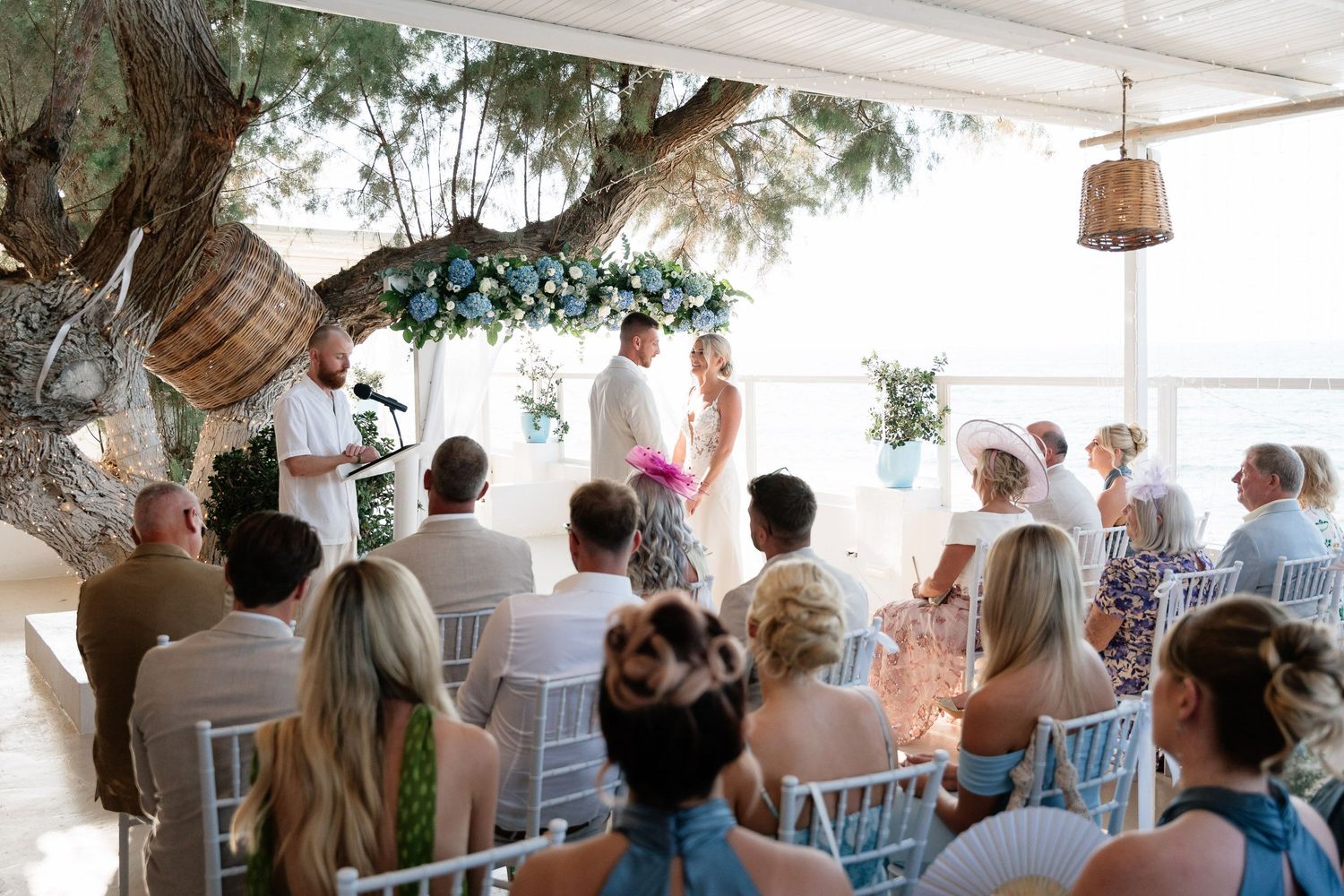 Wedding guests seated at an outdoor reception venue with white decor and natural greenery overhead.