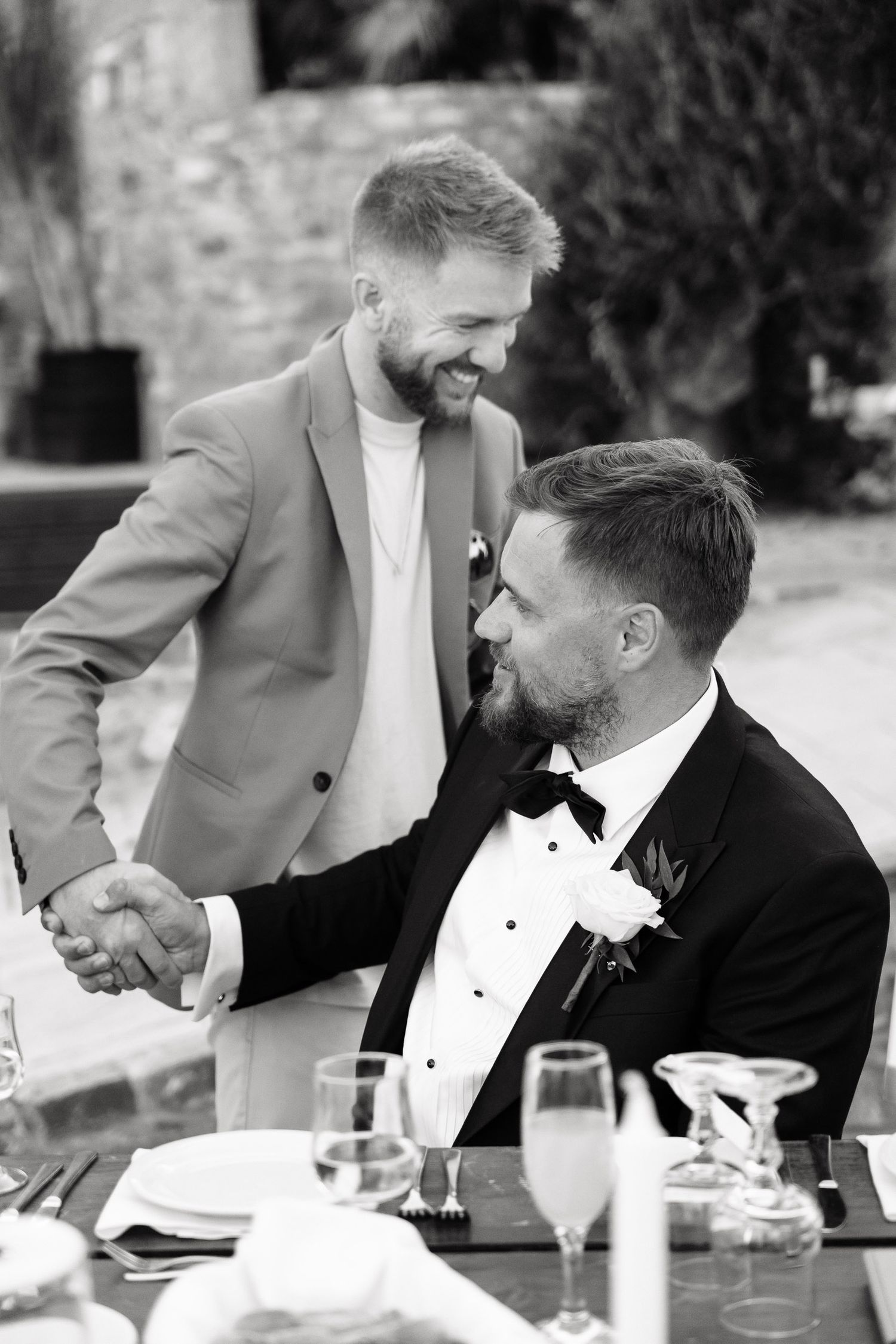Two men in formal attire share a candid moment of laughter while seated at an outdoor dining table.