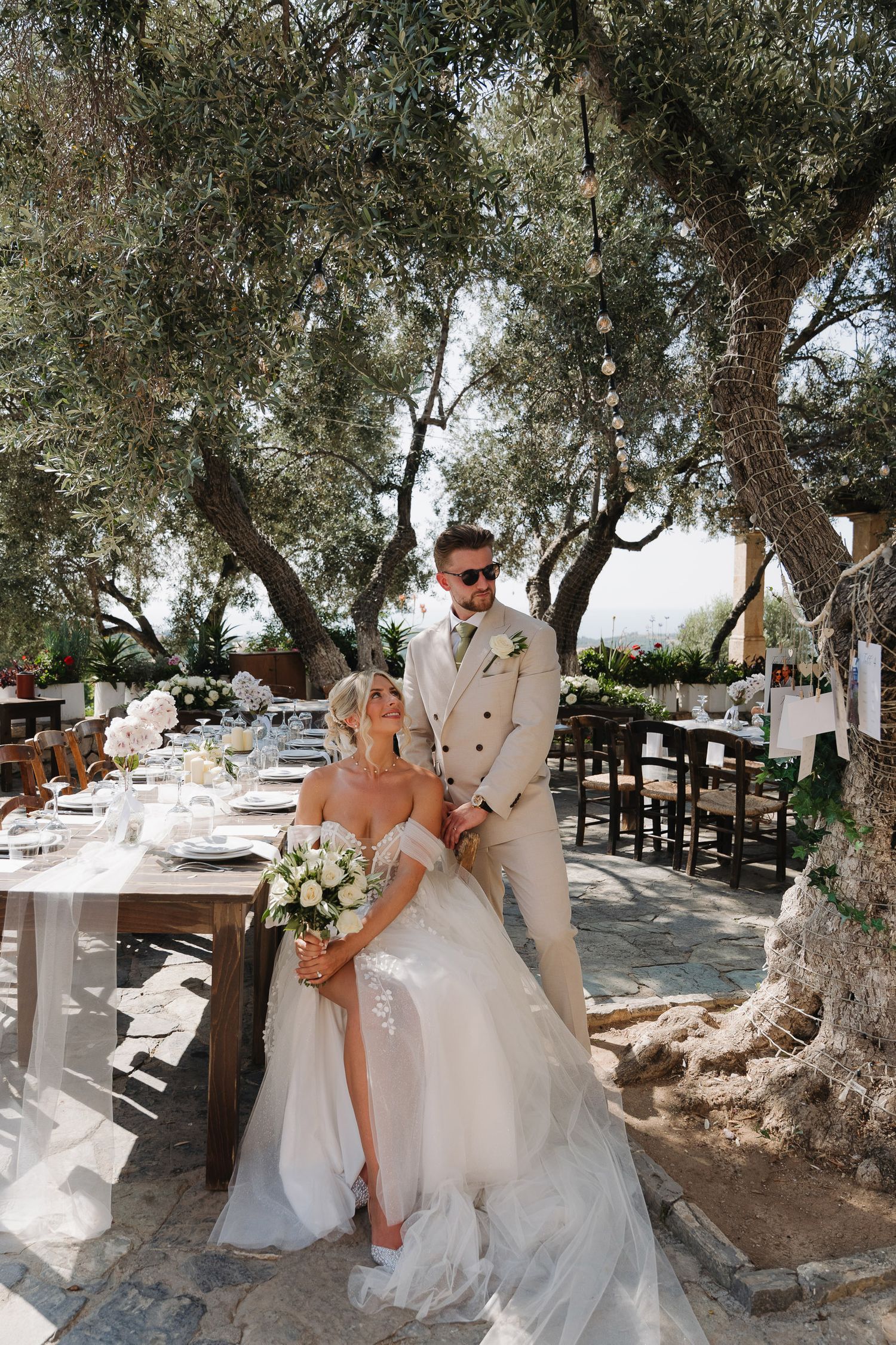 A romantic outdoor wedding scene under olive trees with a bride and groom sharing intimate moments in a Mediterranean setting.