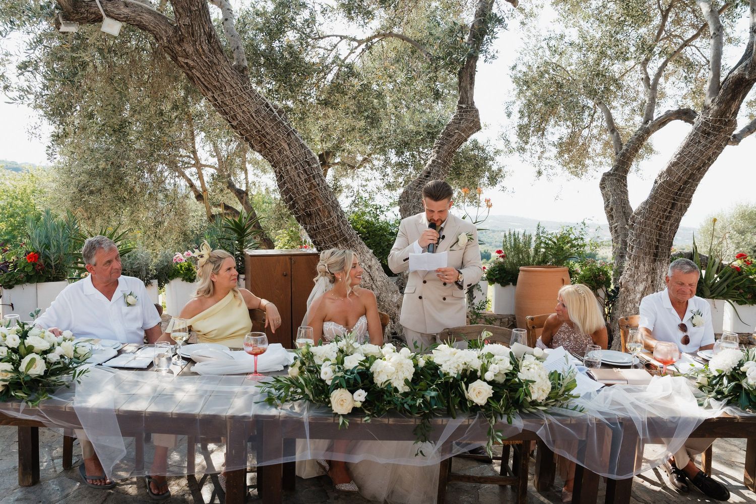 Wedding guests seated at an elegant outdoor reception table under olive trees with white floral centerpieces.
