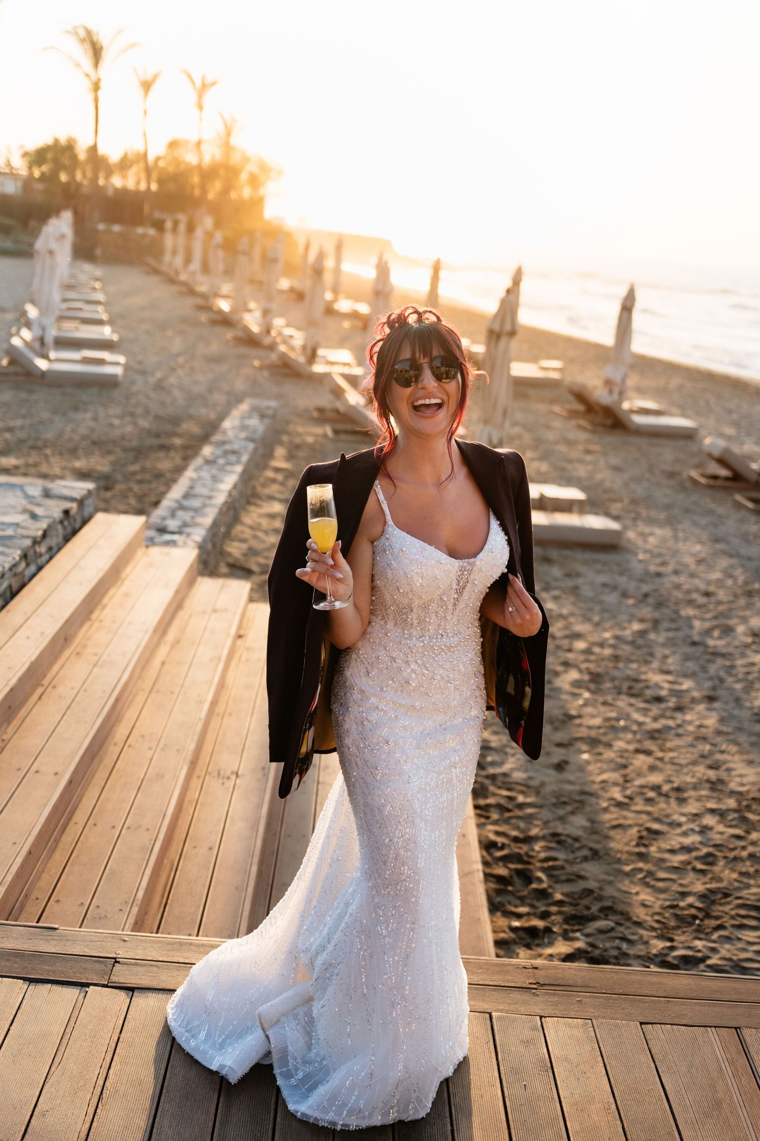 Bride in white lace gown and black jacket holding champagne on beach boardwalk at golden sunset.
