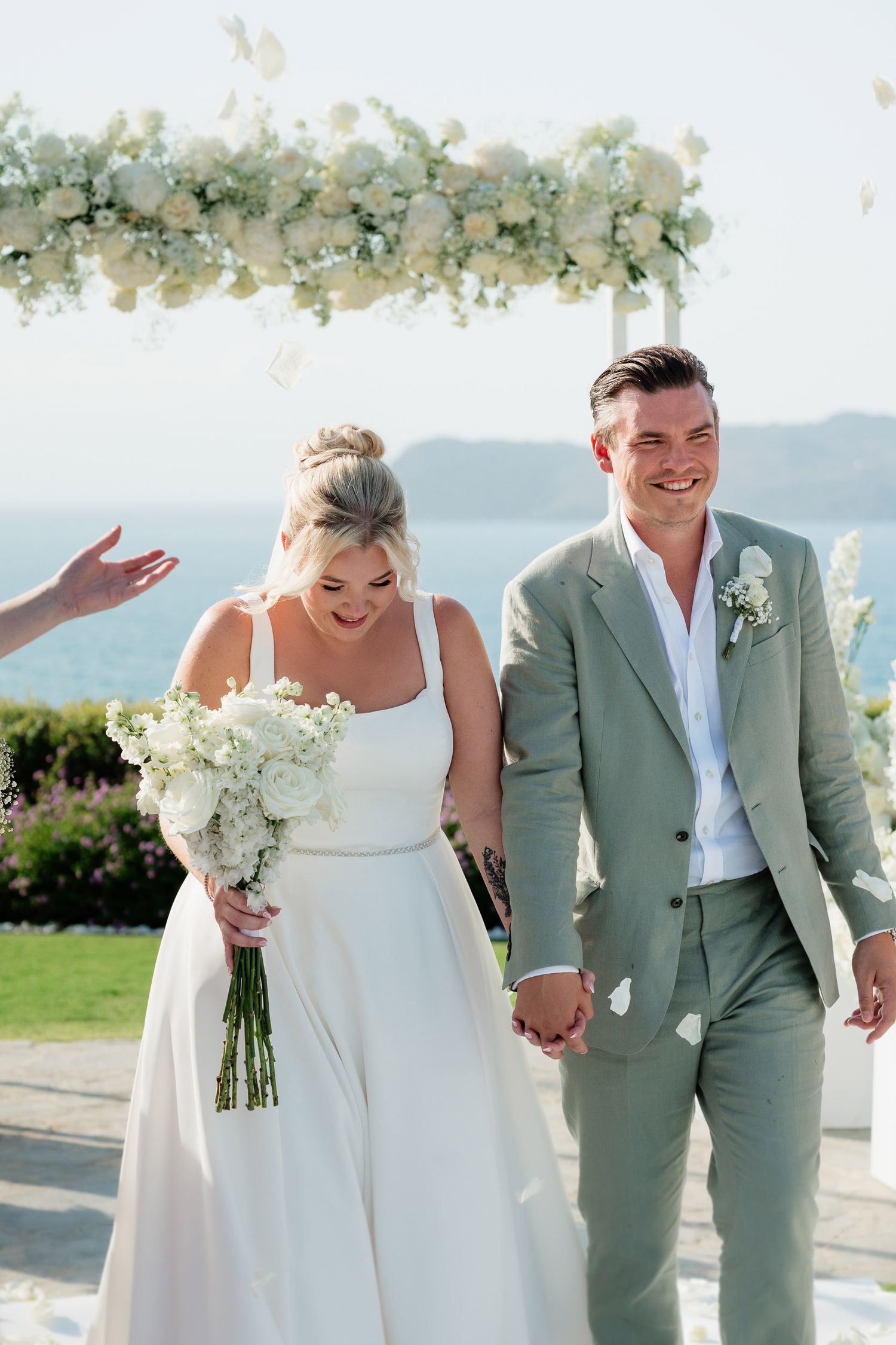 A joyful wedding couple shares candid moments on a sunny waterfront terrace decorated with floral arrangements.