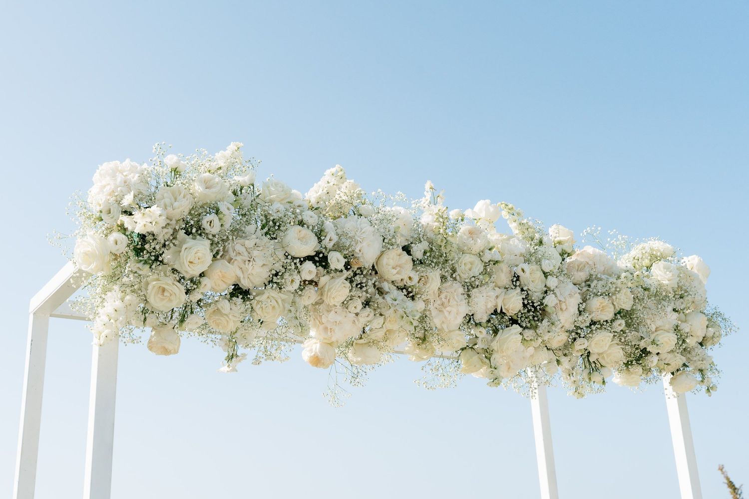 White floral arch decorations against a bright blue sky create an elegant outdoor wedding ceremony setting.