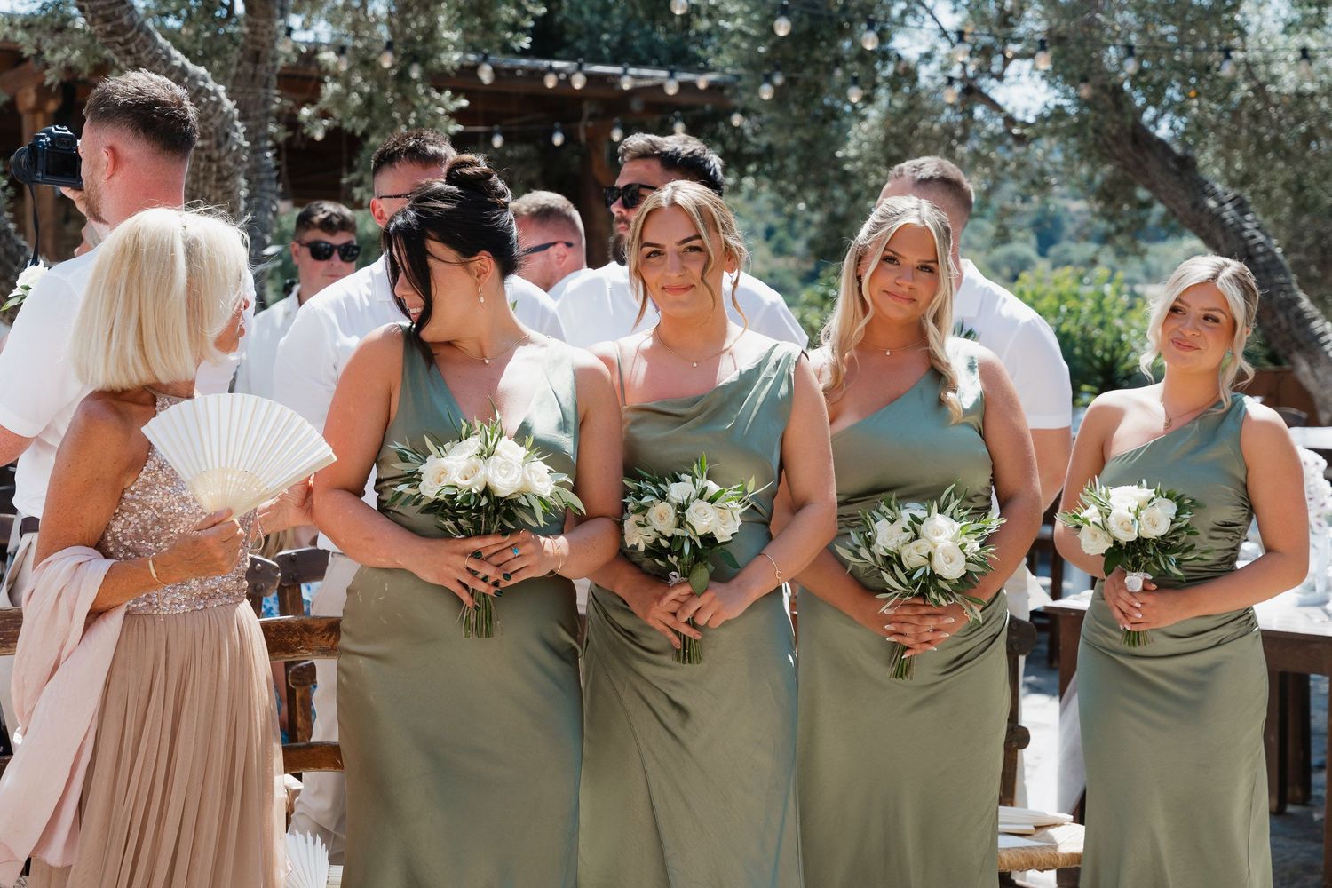 Wedding party members in neutral earth-toned dresses and suits pose together outdoors during a celebration.