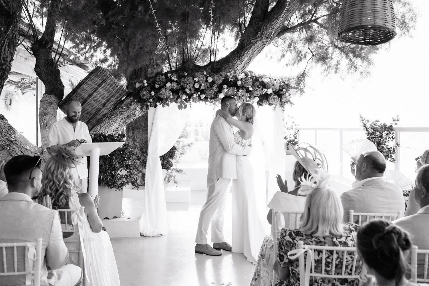A romantic outdoor wedding ceremony under trees with hanging lanterns and guests seated on white chairs.