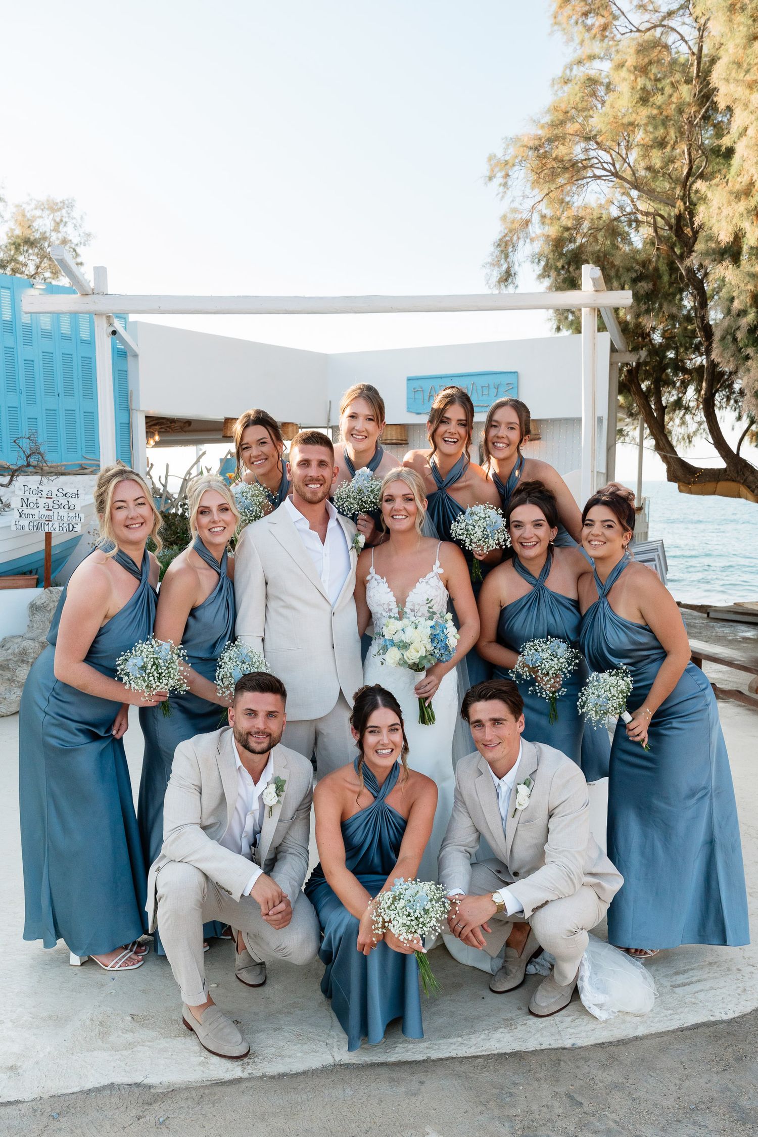 Wedding party in blue bridesmaid dresses and light colored suits poses together on white steps by the ocean.