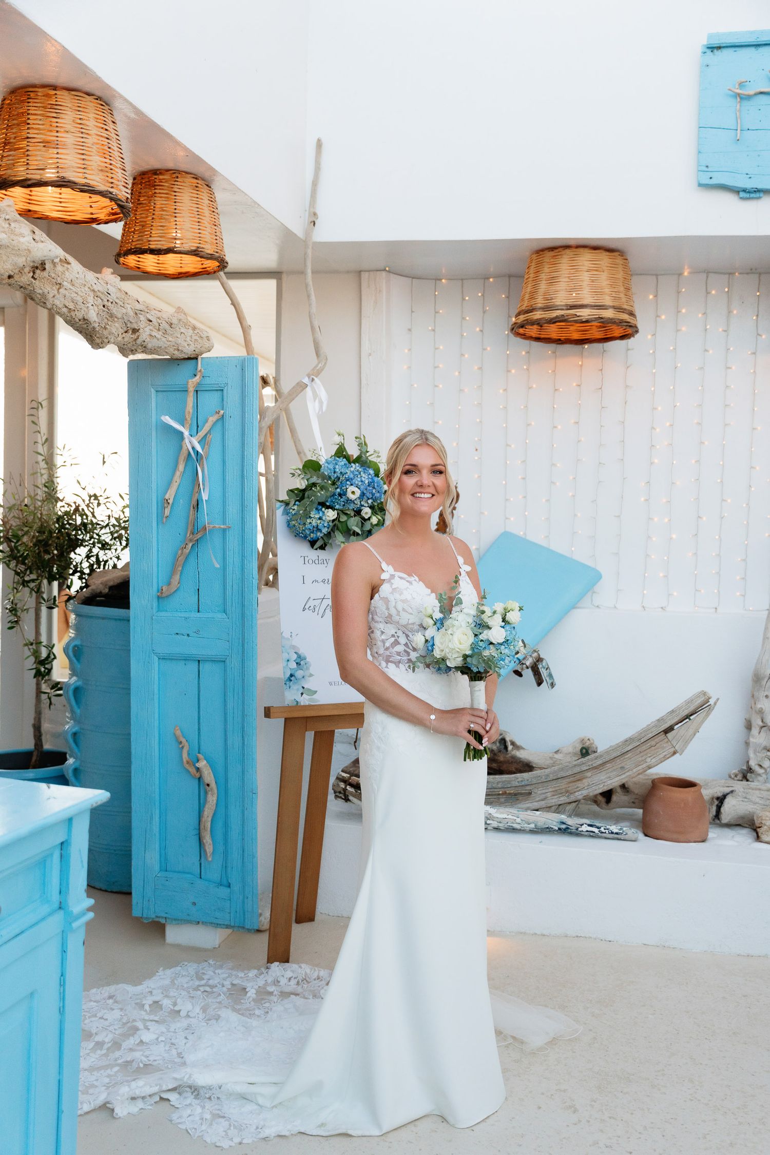 A bride in a sleek white dress holds a bouquet in a Mediterranean-style room with blue accents and wicker pendant lights.