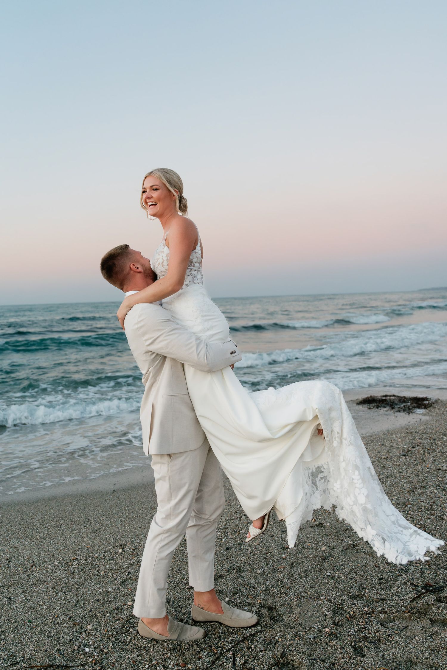 A groom in white lifts and spins his bride on a rocky beach shore with waves crashing in the background.