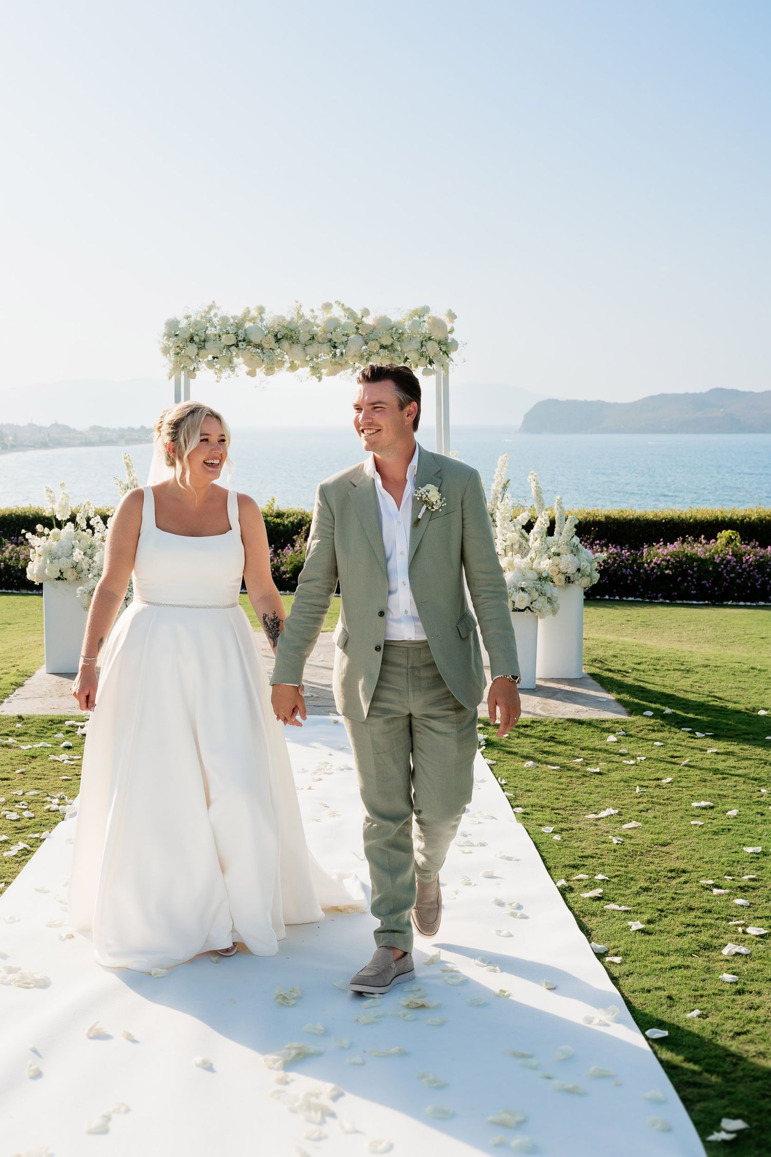 A couple walks down a white aisle runner with scattered flower petals at an outdoor wedding venue overlooking the ocean.