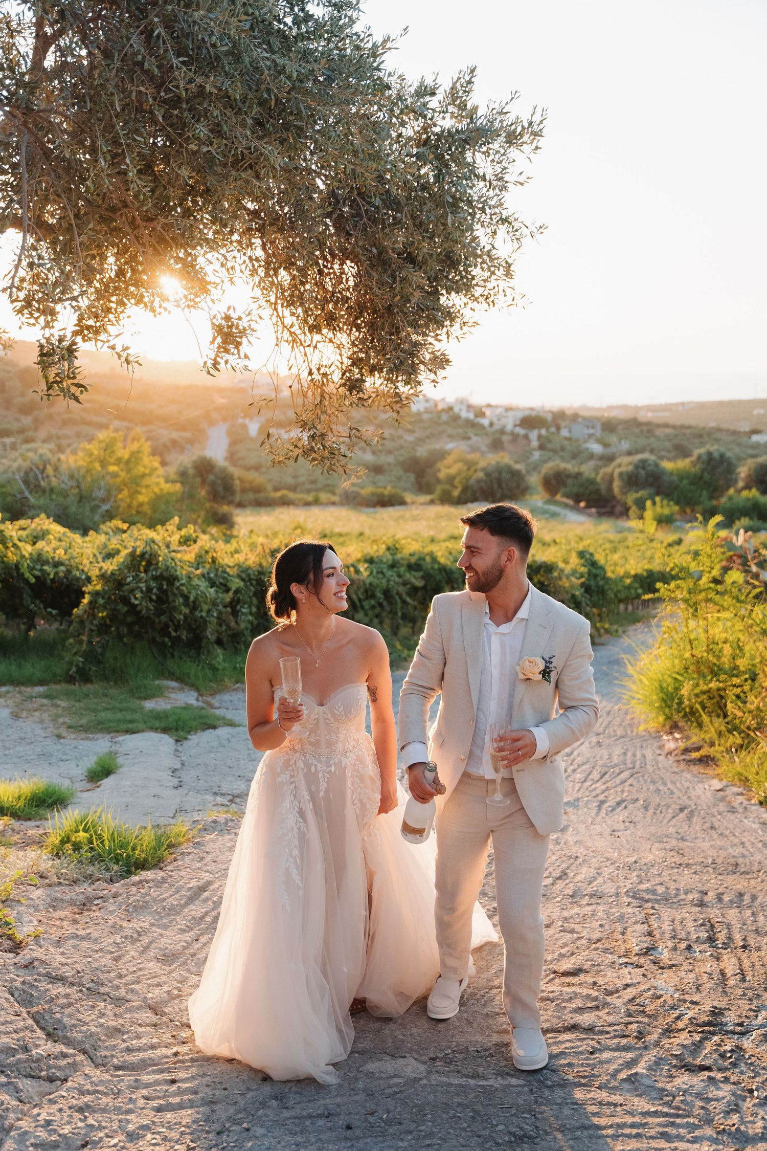 Newlywed couple walks down scenic country path at golden sunset among olive trees and rolling hills in Mediterranean setting.