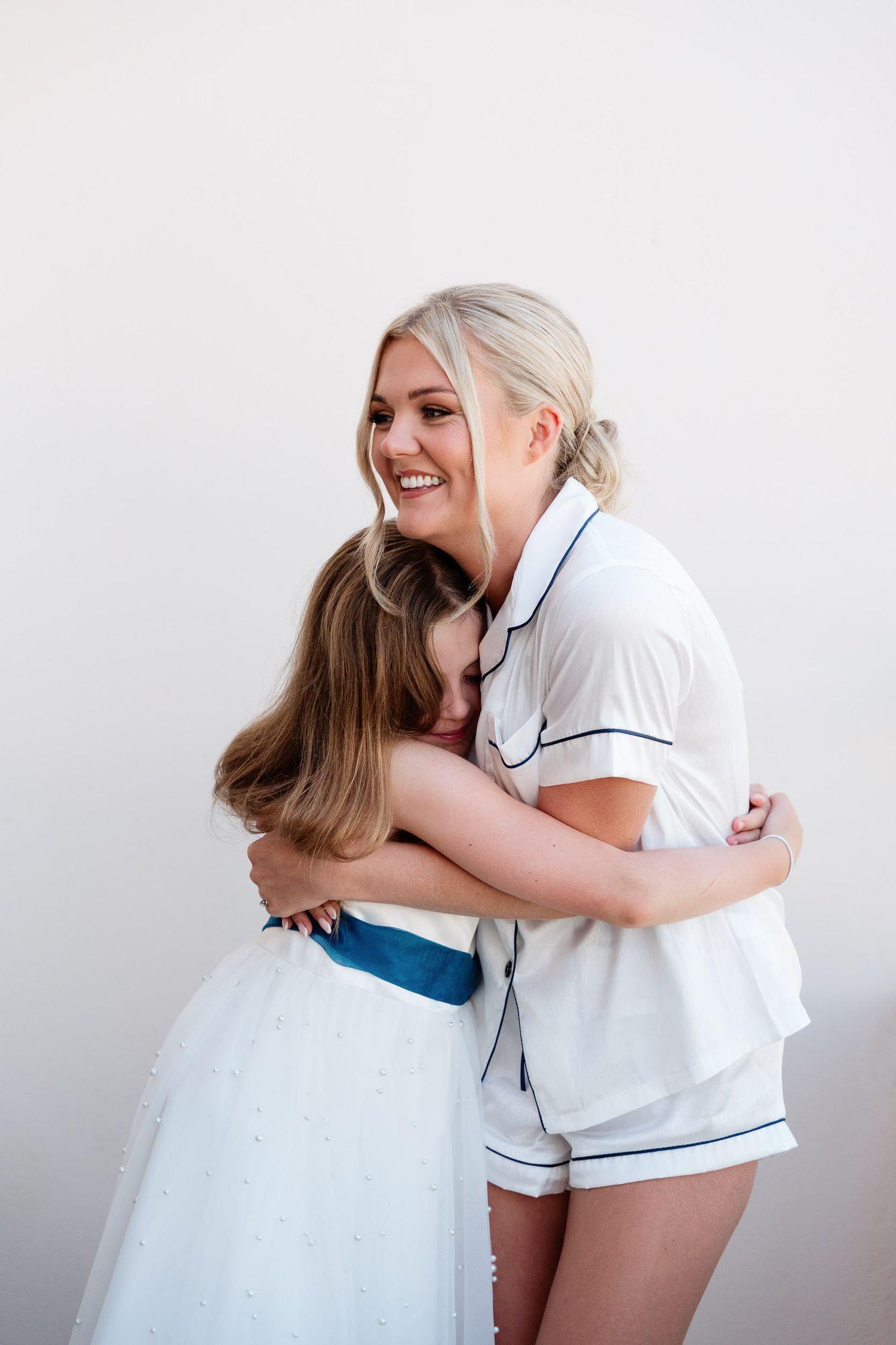 Two people share a warm embrace while wearing white outfits, one in pajamas and one in a dress with blue sash.