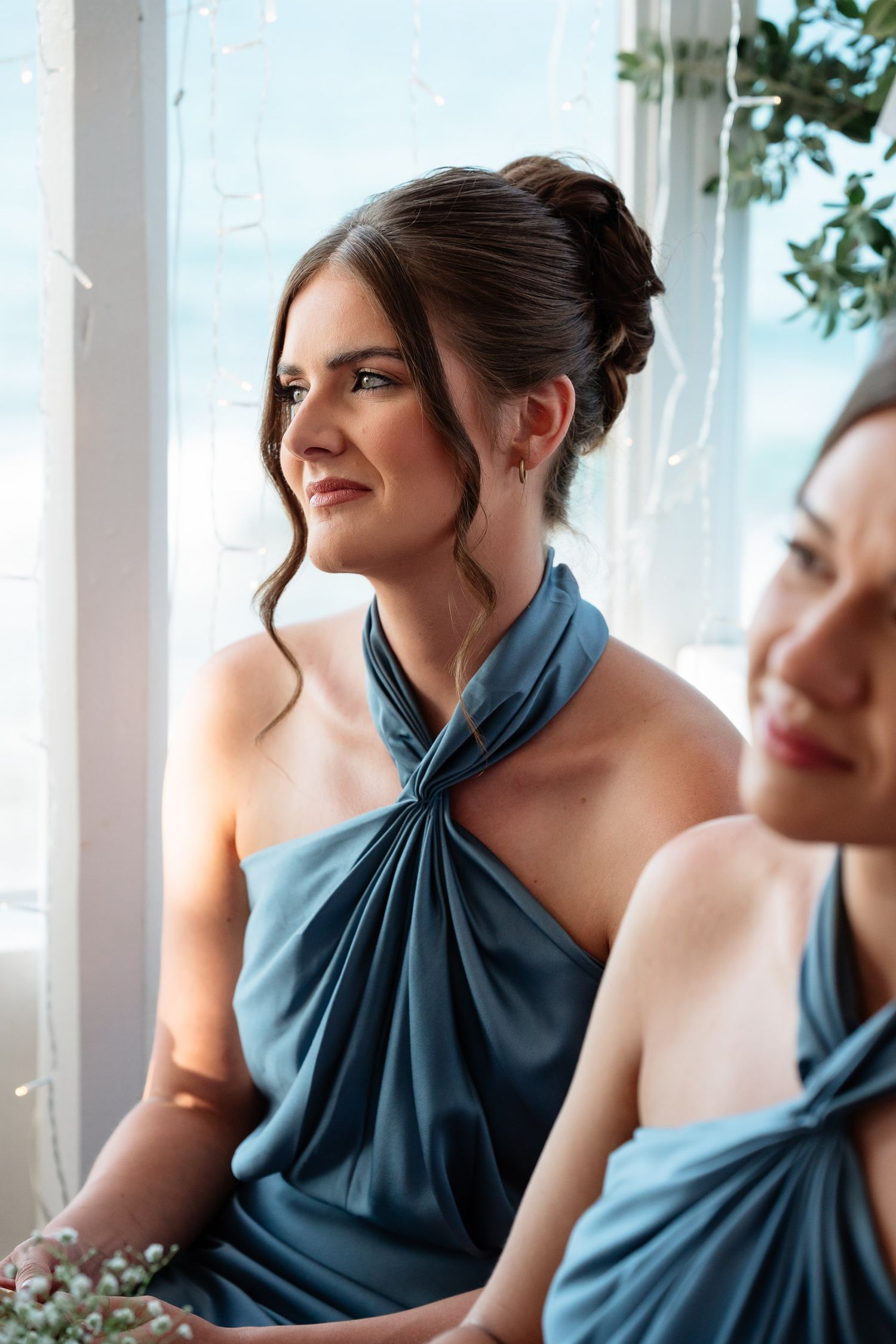 A bridesmaid in an elegant dusty blue halter dress smiles while holding a bouquet during a wedding celebration.