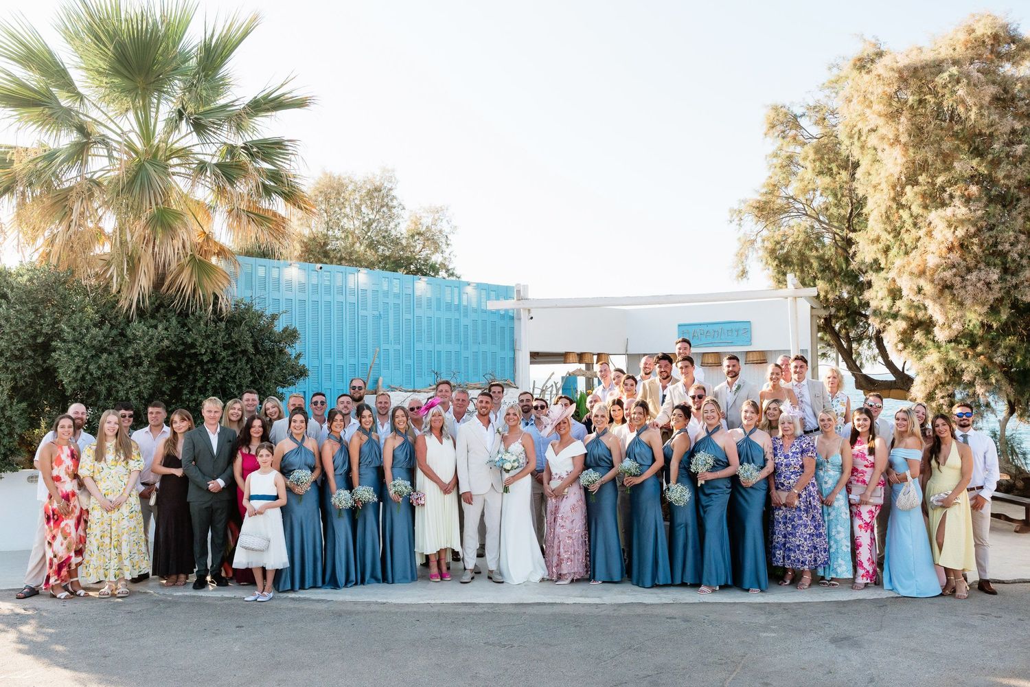 Large wedding party in coordinated blue and white attire poses together outdoors for group photo at sunset.