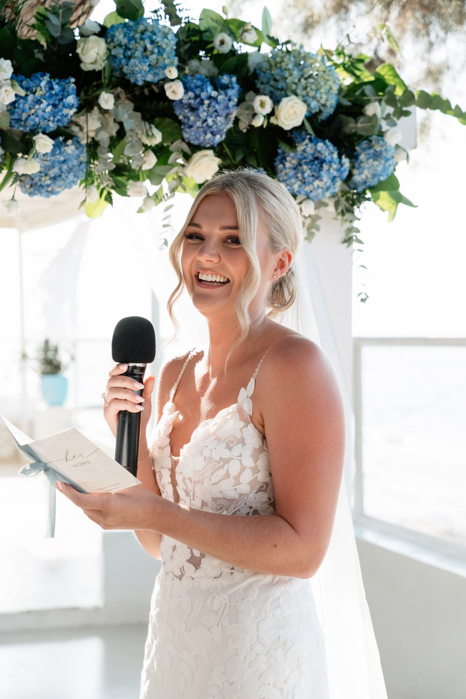 A bride in a white lace gown laughs while holding a phone under blue hydrangea floral arrangements.