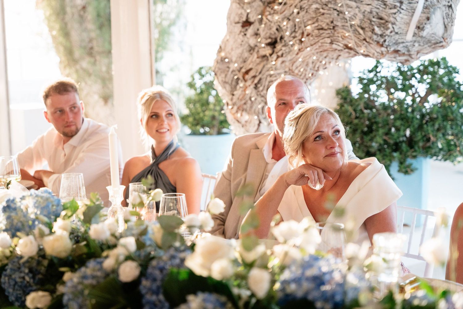 Wedding guests seated at an elegant outdoor reception with floral centerpieces and white decor under a covered venue.