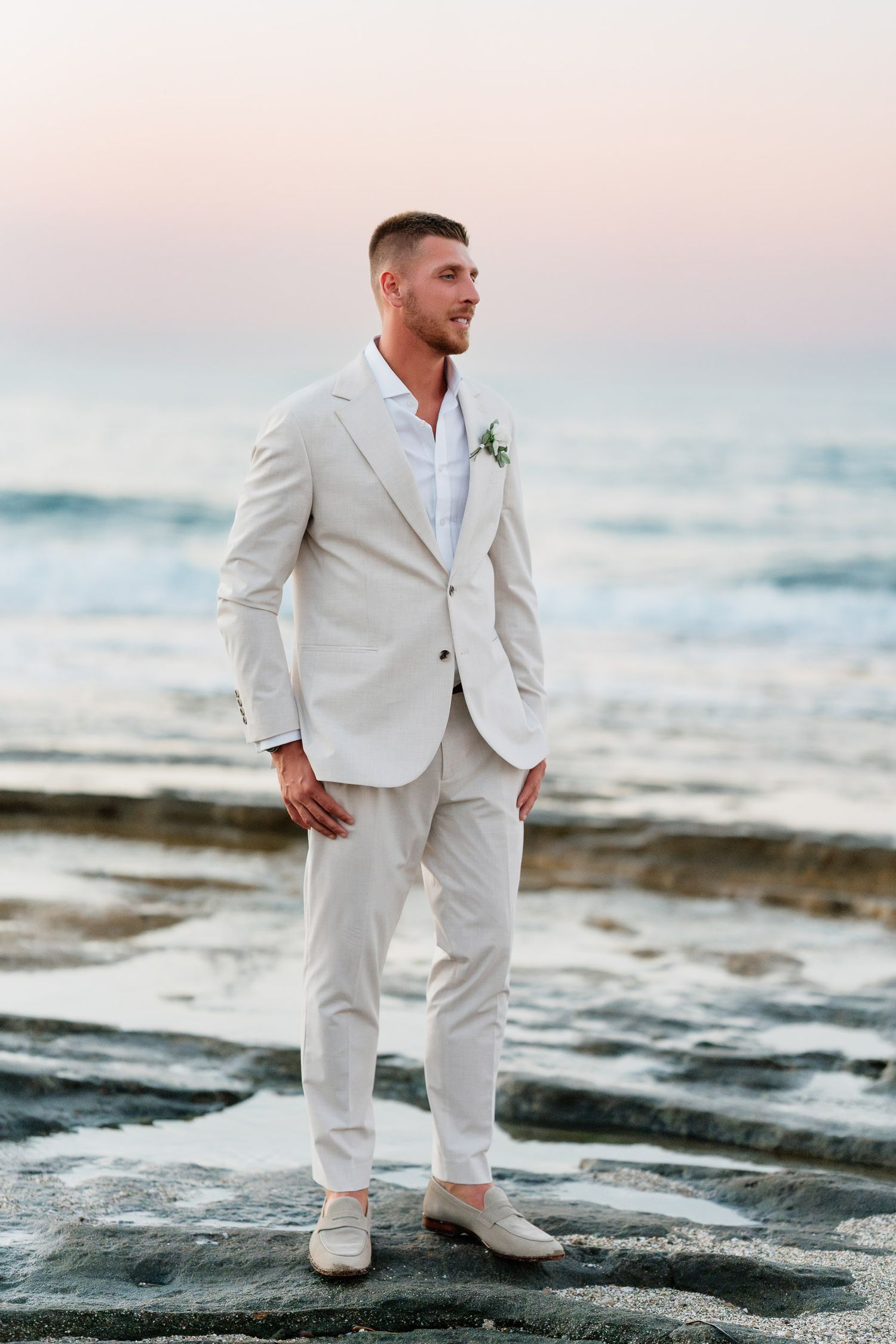 Groom in crisp white suit poses on sandy beach during sunset with ocean waves in background.