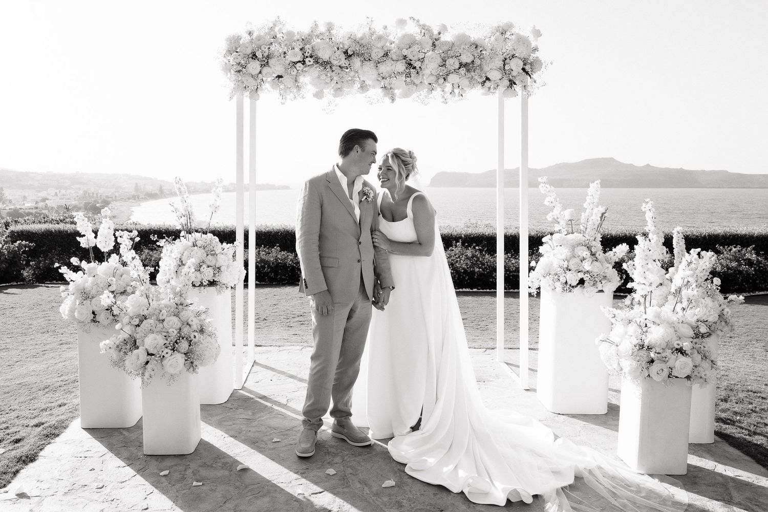 A romantic outdoor wedding ceremony setup with white floral arrangements and flowing fabric draping under a white pergola.