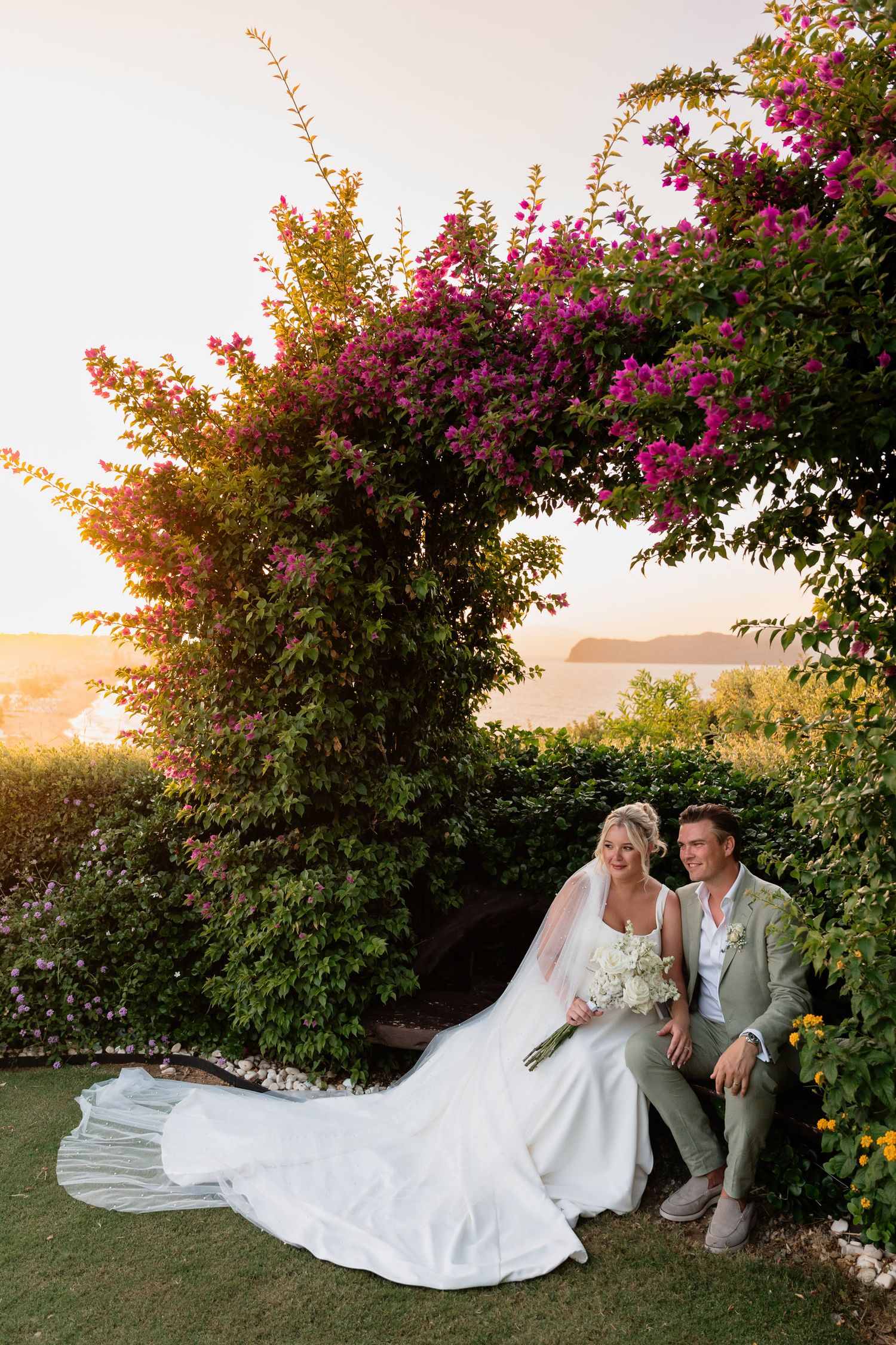 A couple sits under a flowering bougainvillea arch during sunset at an outdoor garden wedding ceremony.