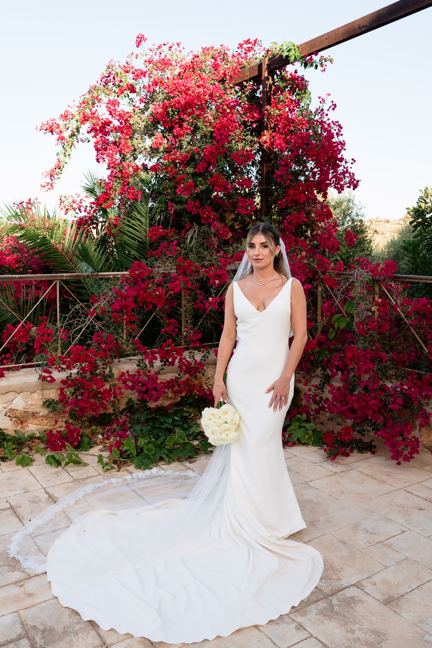 A white wedding dress flows elegantly against vibrant pink bougainvillea flowers in a Mediterranean garden setting.