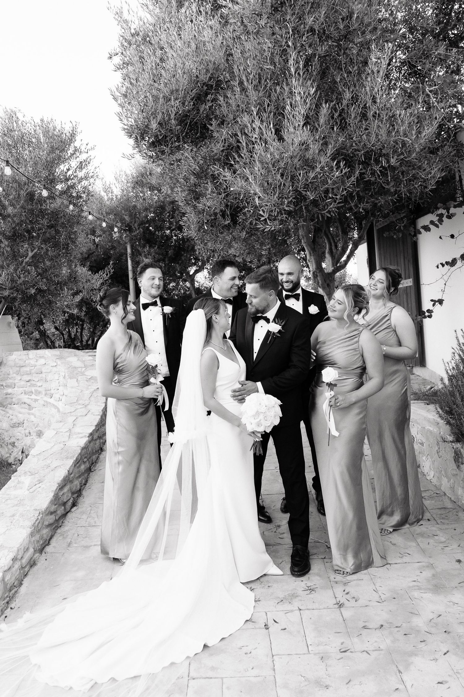 Wedding party poses together on a sandy beach path surrounded by lush greenery and trees in black and white photography.