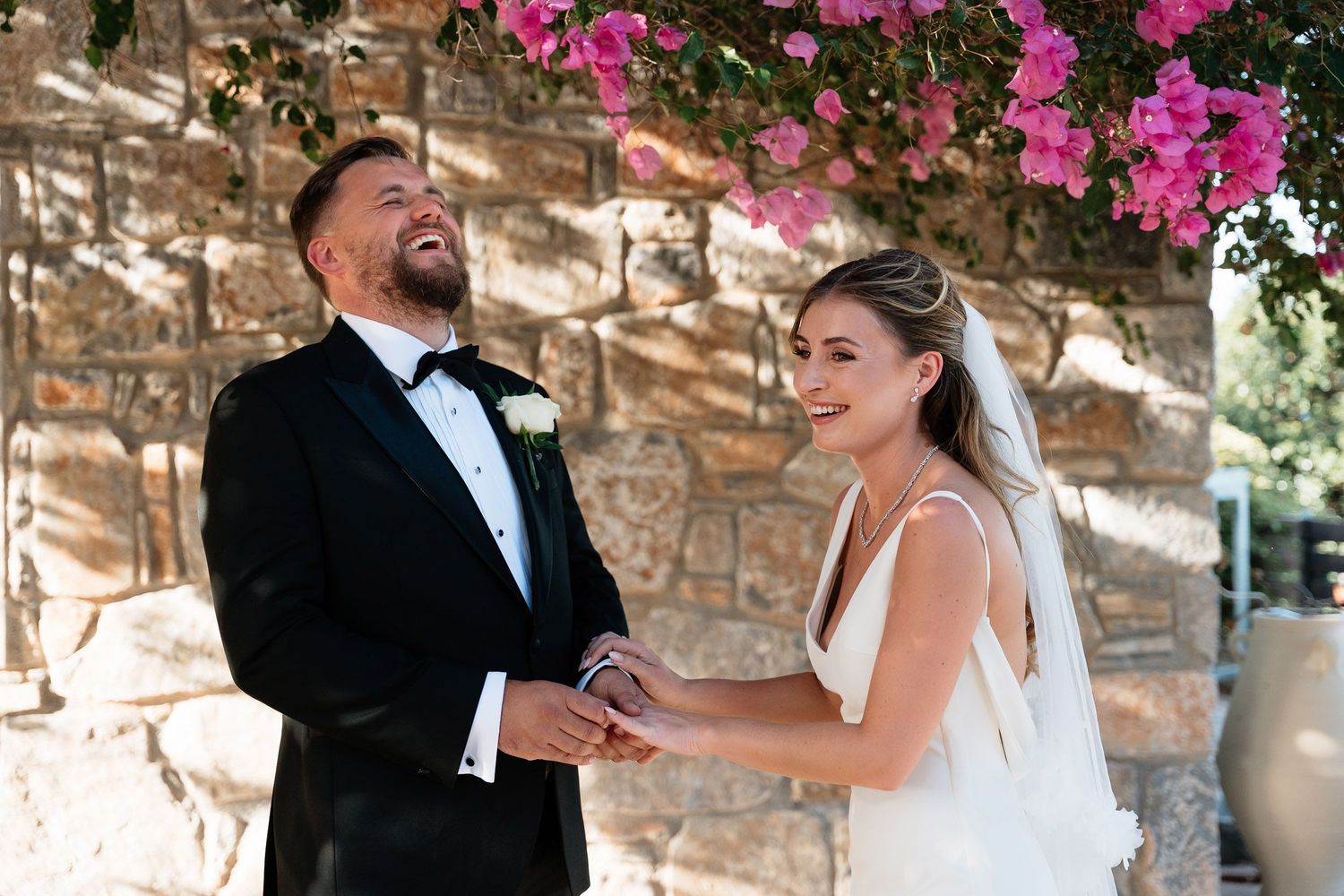 A joyful couple shares a tender moment during their wedding ceremony under a stone archway adorned with pink flowers.