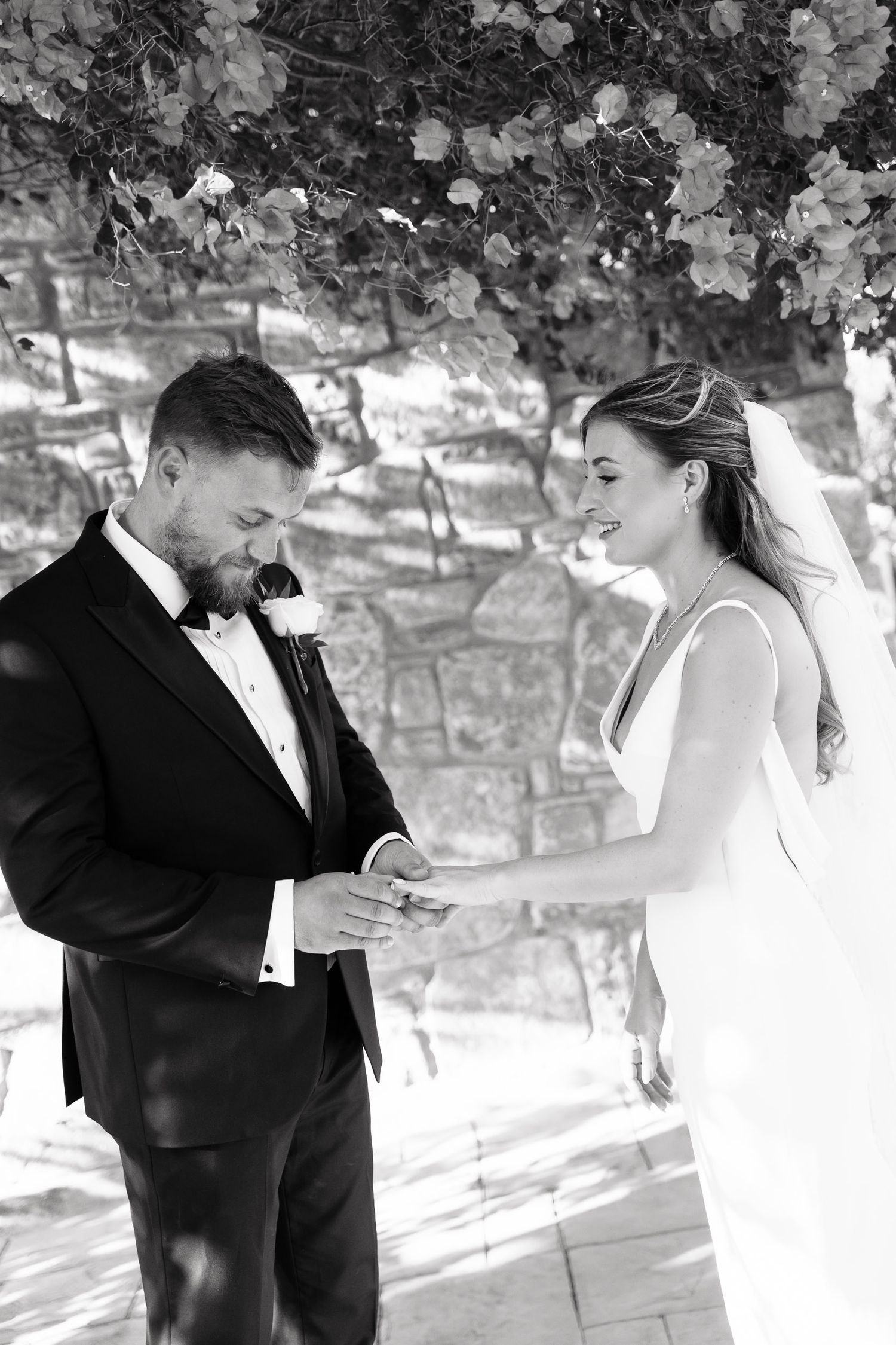 A black and white wedding photo showing a couple during their ceremony beneath flowering trees against a stone wall.