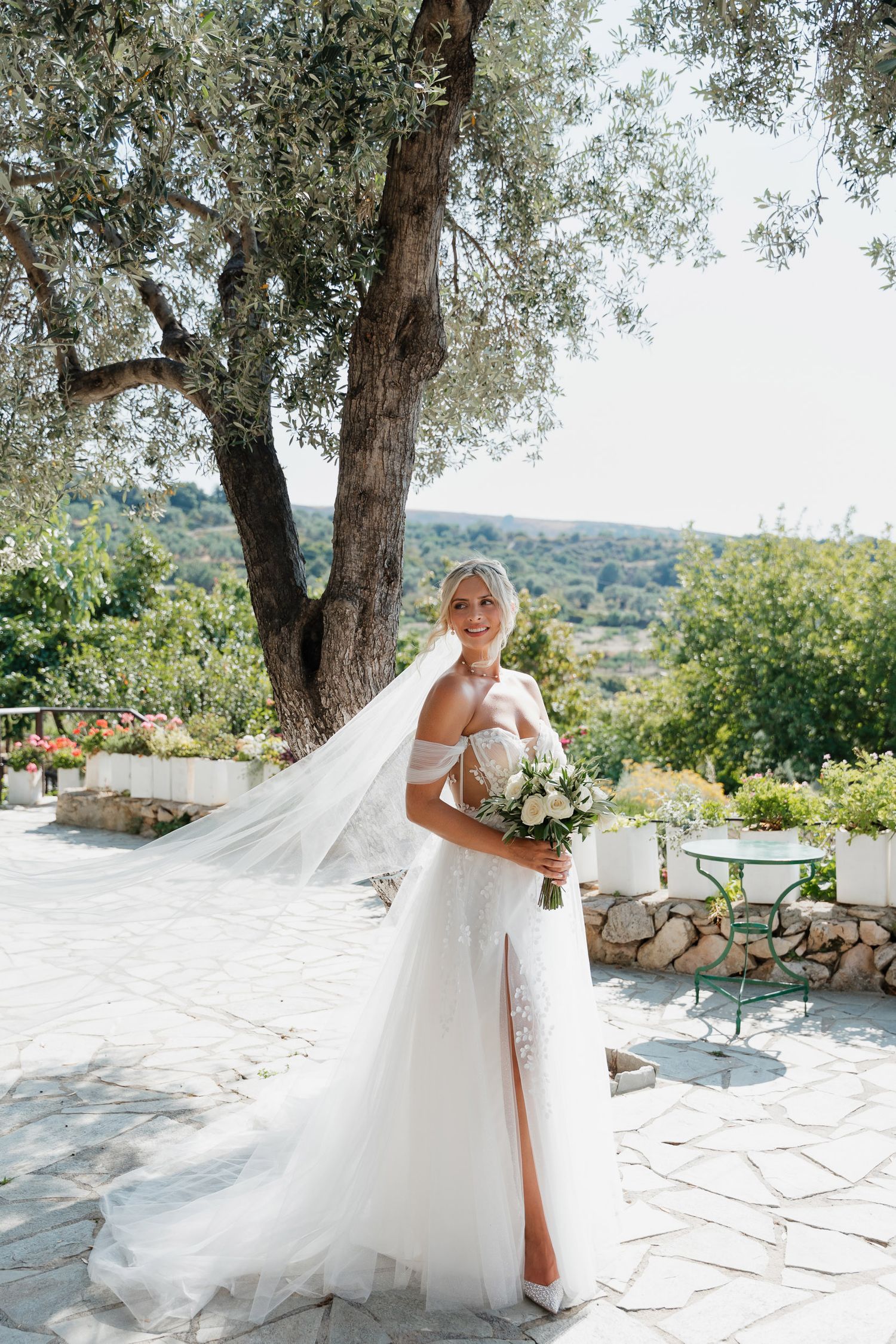 A flowing white wedding dress with a long veil billows in the breeze against a Mediterranean landscape with olive trees.