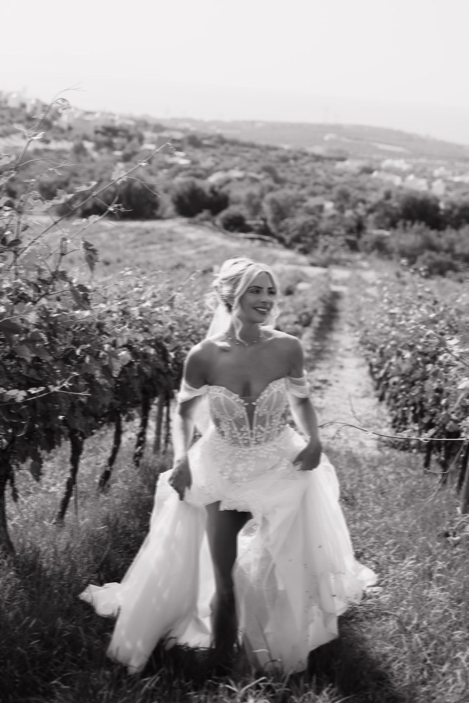A black and white photo shows a bride in a strapless wedding gown walking along a scenic hillside path through nature.