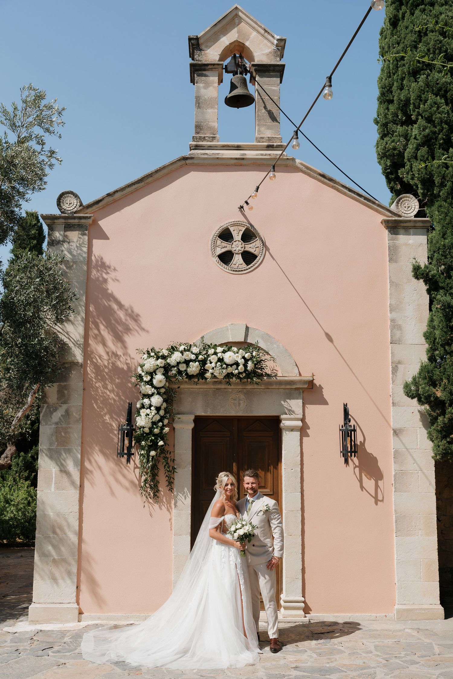 A romantic wedding photoshoot outside a charming pink Mediterranean chapel with white floral arch decorations.