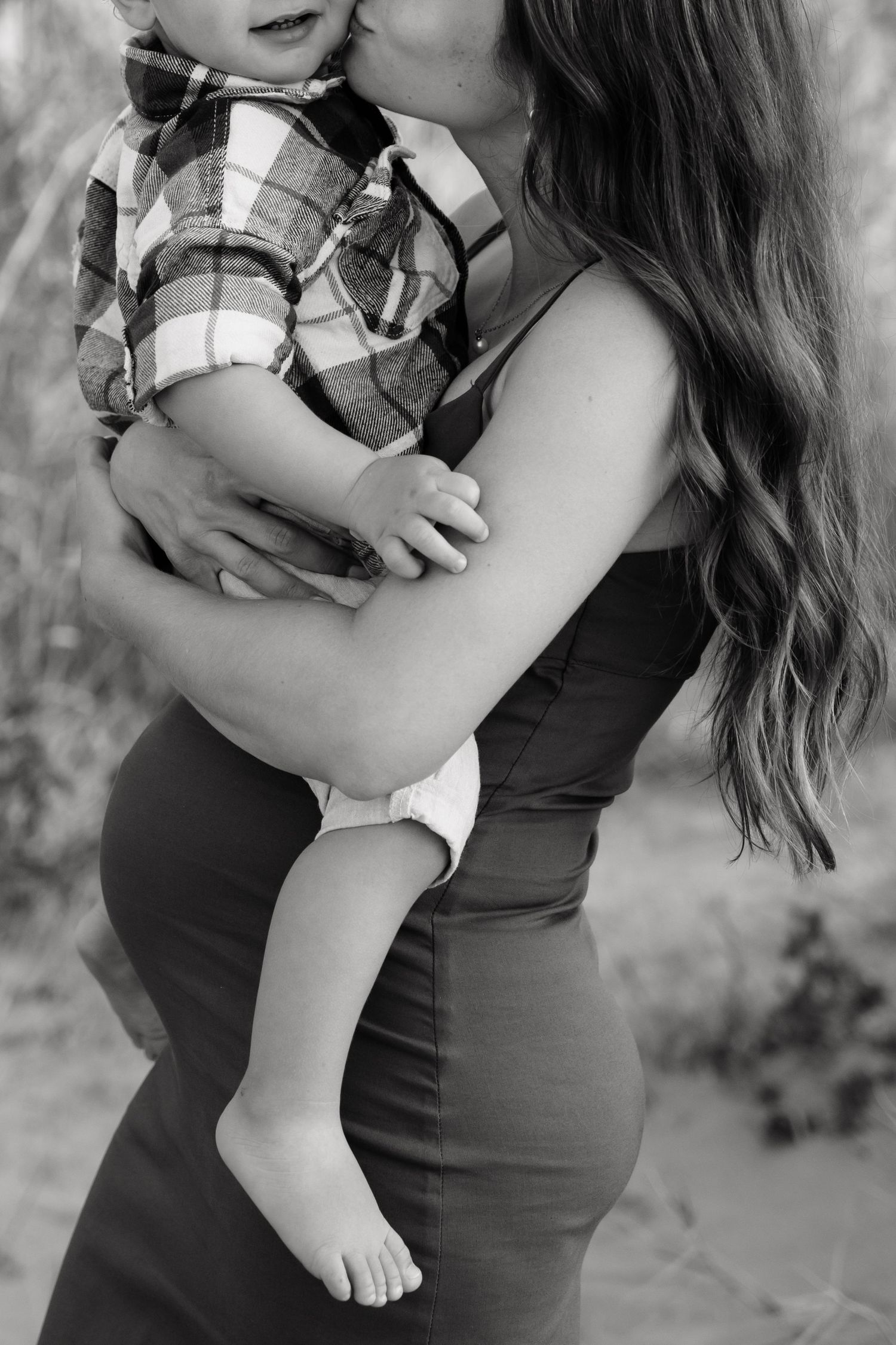 Black and white maternity photo series showing woman in dark dress holding and embracing small child in plaid shirt.