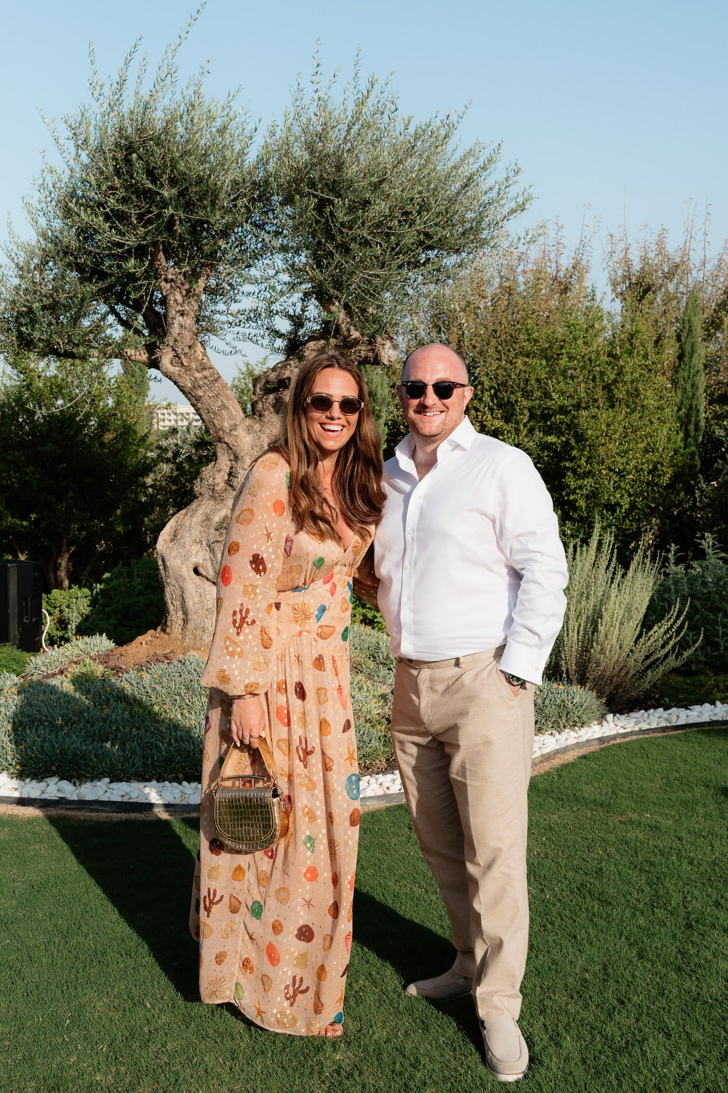 A couple poses together outdoors on a sunny day with olive trees and greenery in the background.