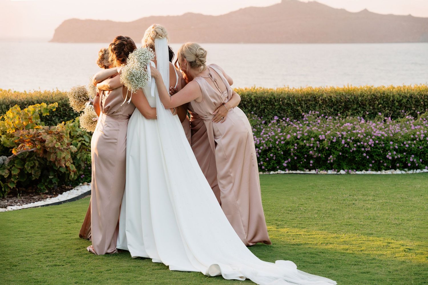 Bridesmaids in blush pink dresses gather around bride in white gown during golden hour at outdoor wedding.