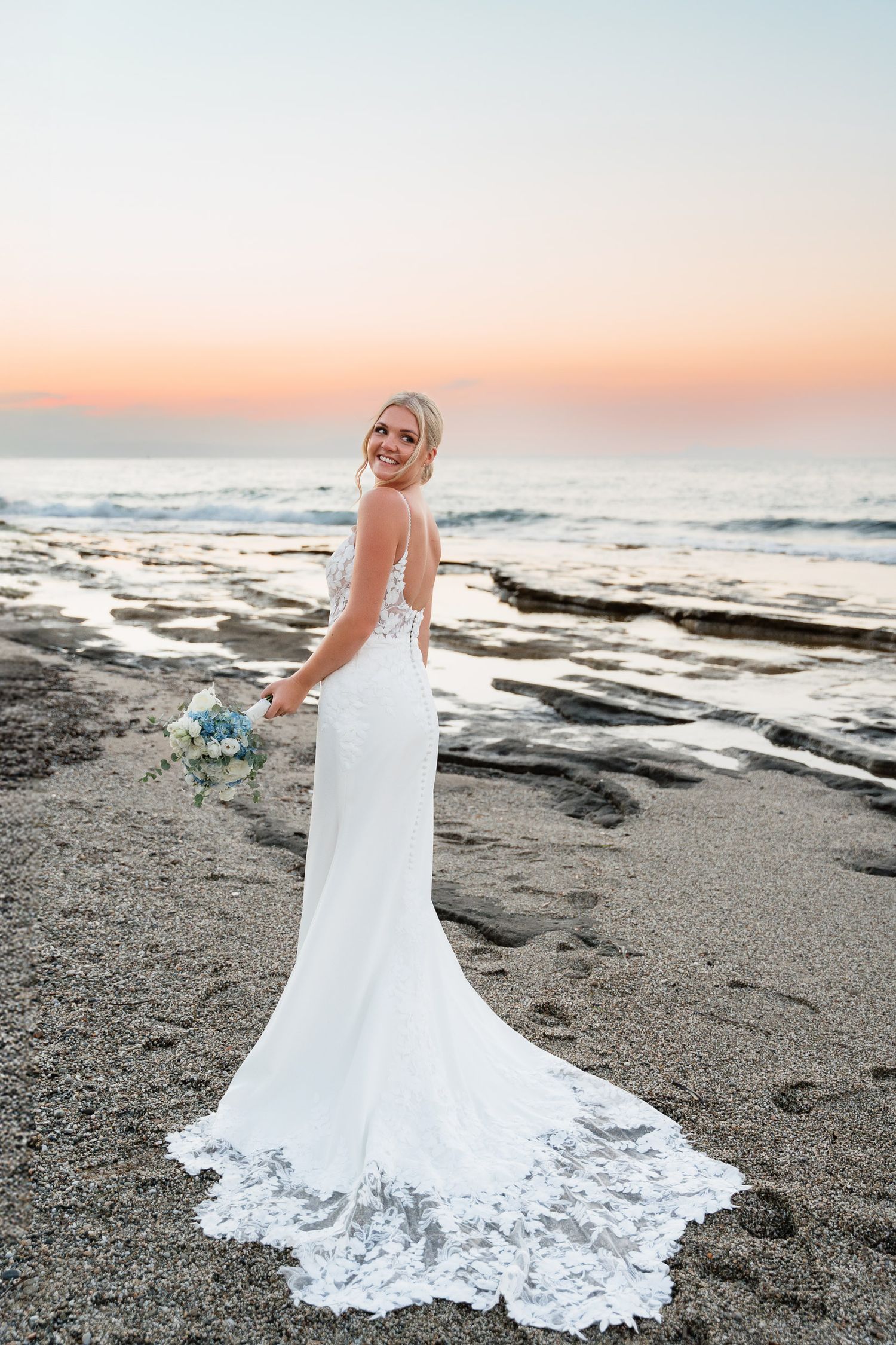 A white wedding dress with lace details trails elegantly across a dark sandy beach at sunset.