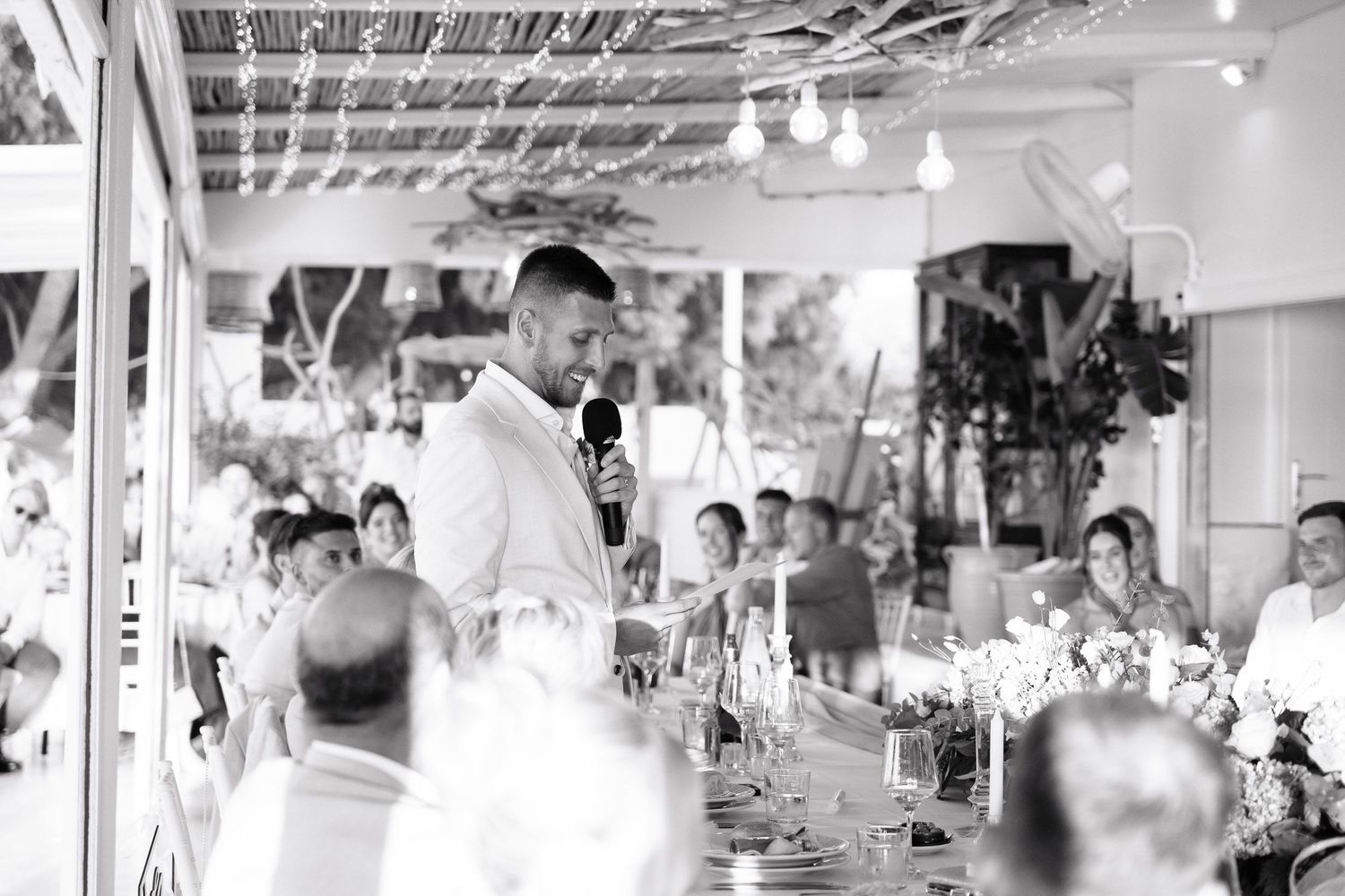 Black and white panoramic photo of a crowded indoor event space with people seated at tables and decorative lighting.