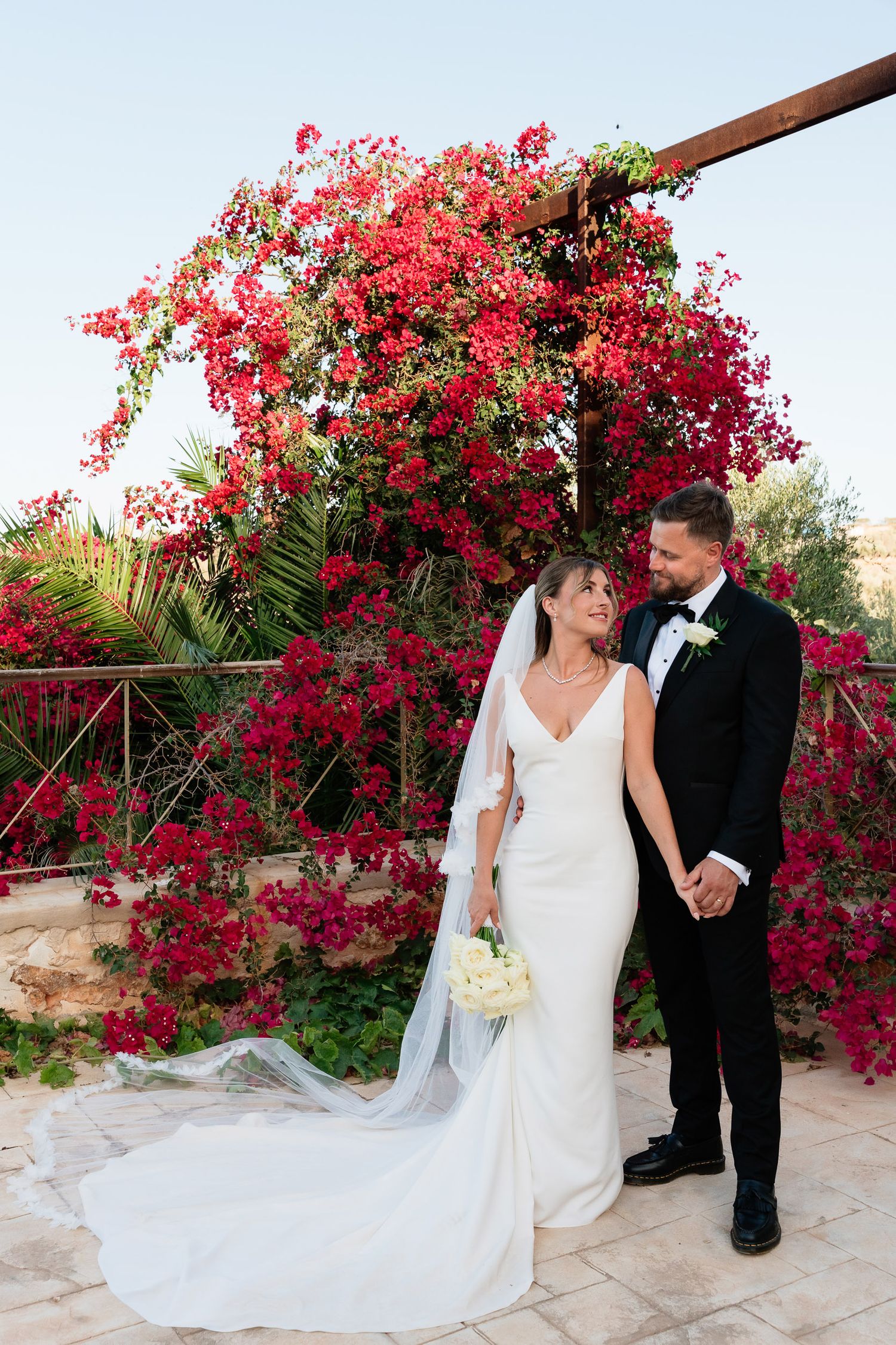 A couple in wedding attire stands together near vibrant red bougainvillea flowers in a garden setting.